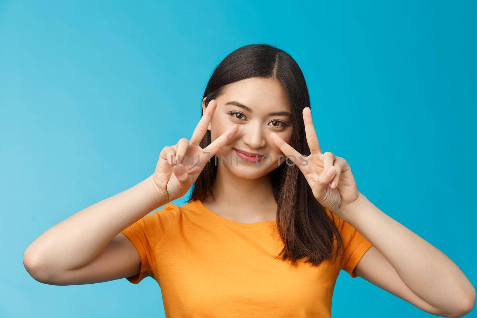 Tender cute asian woman short dark haircut show peace victory signs near cheeks, posing joyfully grinning optimistic, determined win, tilt head coquettish, stand blue background cheerful.