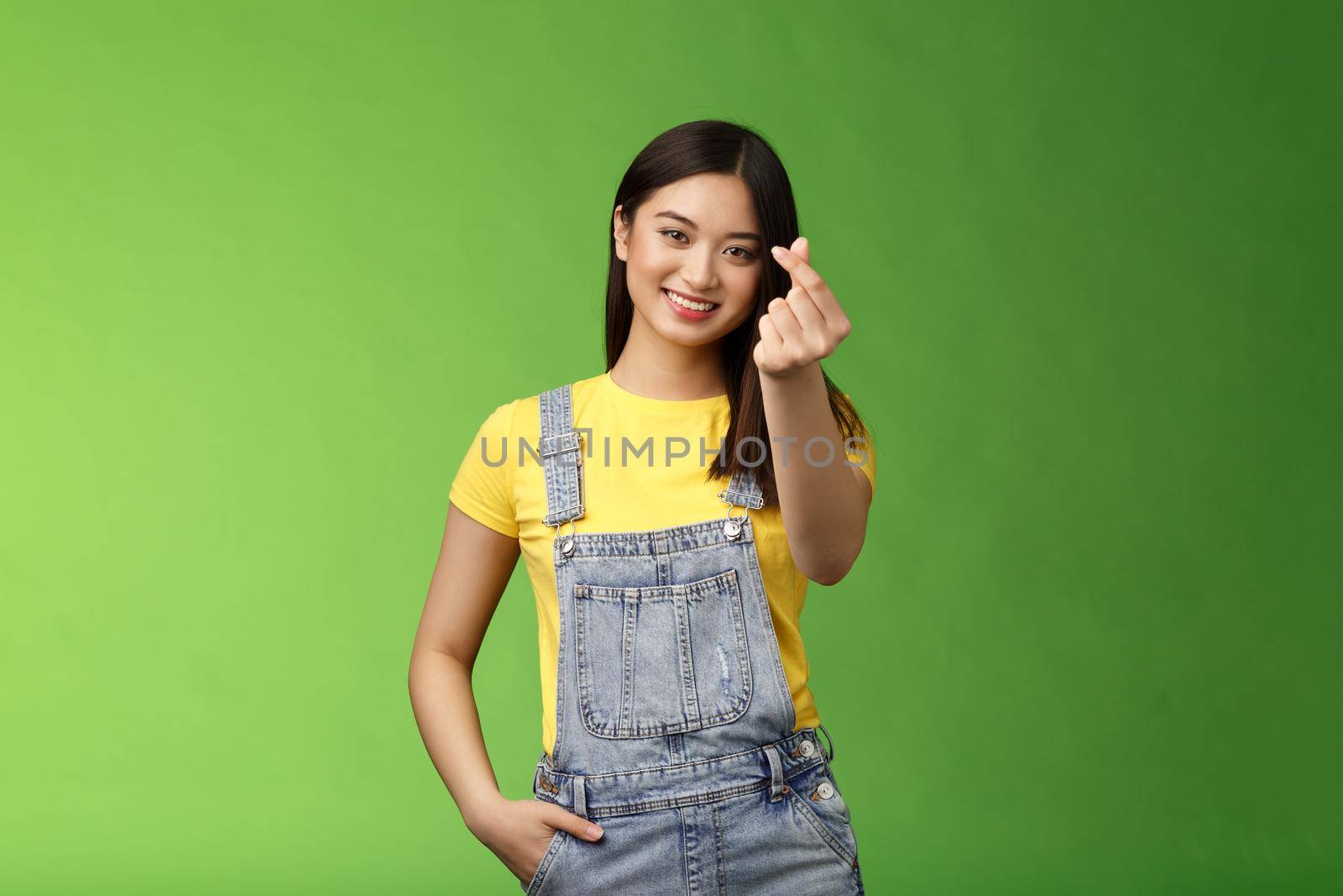 Lovely tender carefree asian girlfriend show korean love sign, make finger heart smiling cute, showing passion and affection, stand green background in overalls and yellow t-shirt, express sympathy by Benzoix