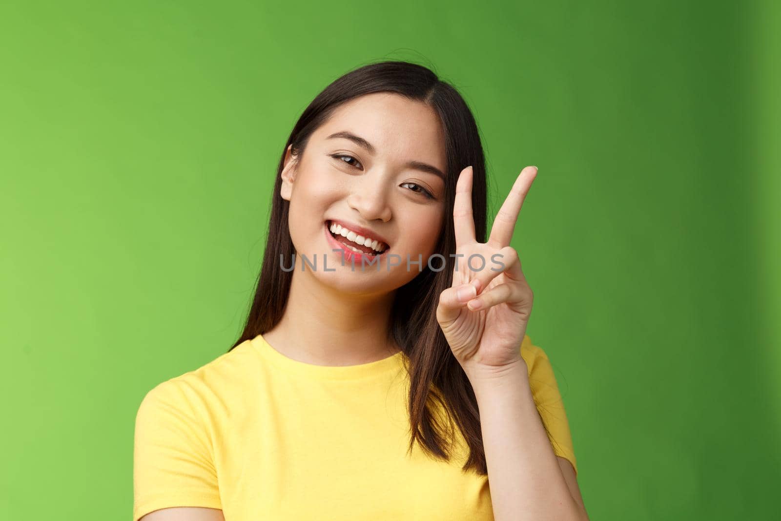 Close-up joyful cute asian girl lively look camera smile, show peace victory sign entertained, grinning posing photograph tender expression, stand green background positive and lucky.