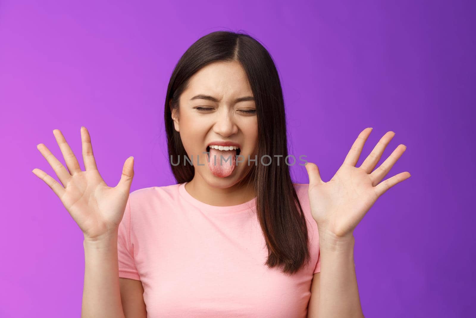 Close-up silly moody asian girl complaining act immature and bothered, raise hands unsatisfied show tongue aversion disgust, close eyes, react terrible disgusting thing, stand purple background.