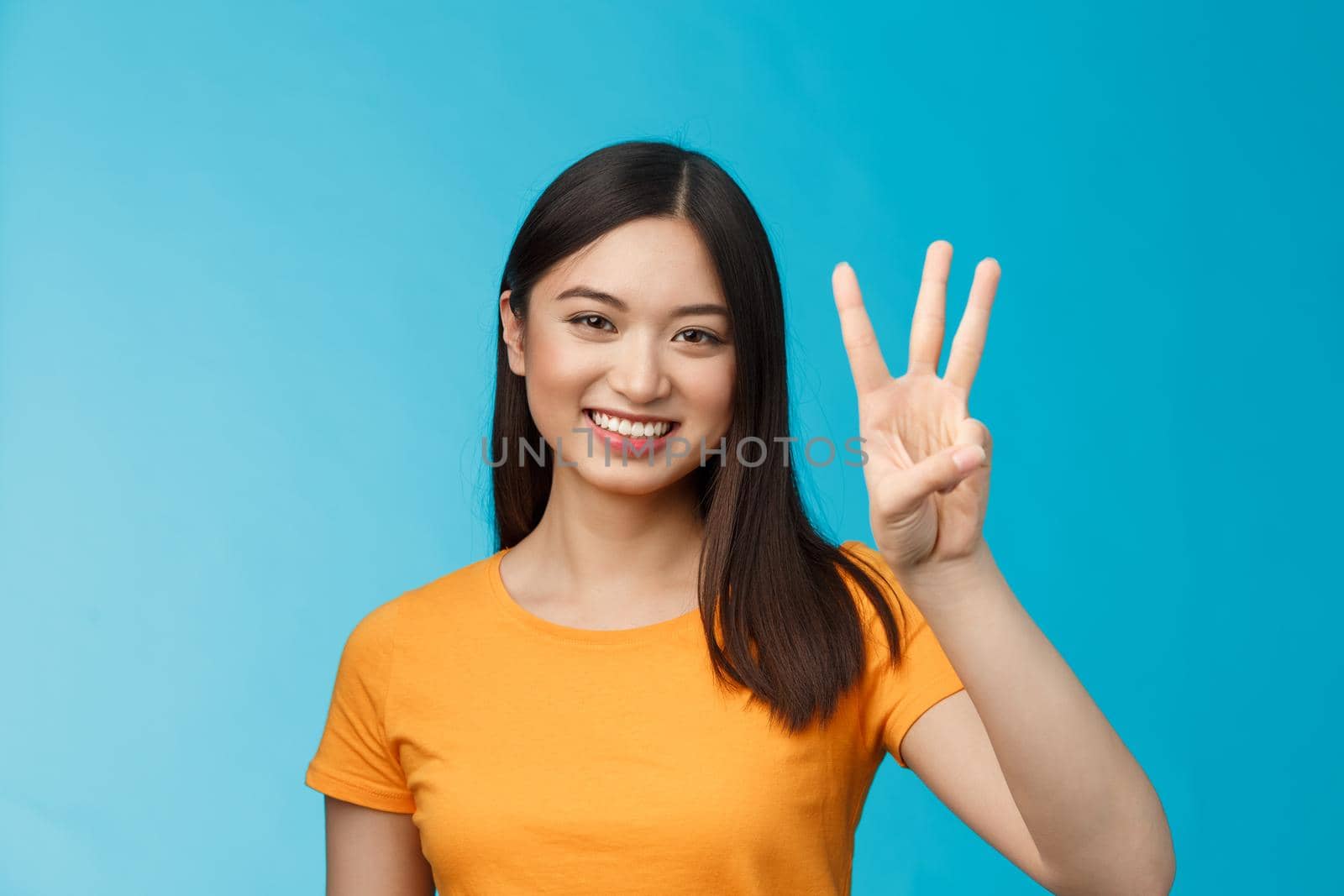 Friendly enthusiastic tender asian woman making reservation for three, number third, smiling broadly, stand blue background joyfully, posing near blue background in yellow t-shirt by Benzoix