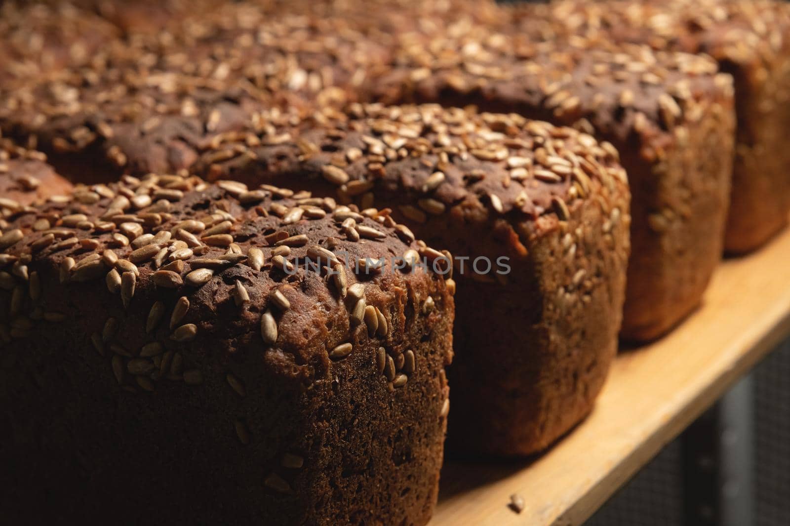 A lot of whole delicious homemade rectangular rye bread with sunflower seeds on top lies on a wooden rack background.