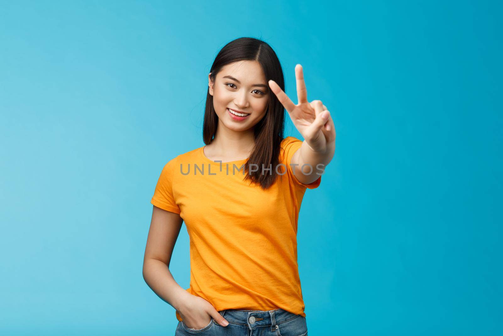 Cheerful friendly asian girl pacifist smiling happily show peace, victory sign, determined win aim achieve success stand blue background joyful, feel luck enthusiastic, wear yellow summer t-shirt.