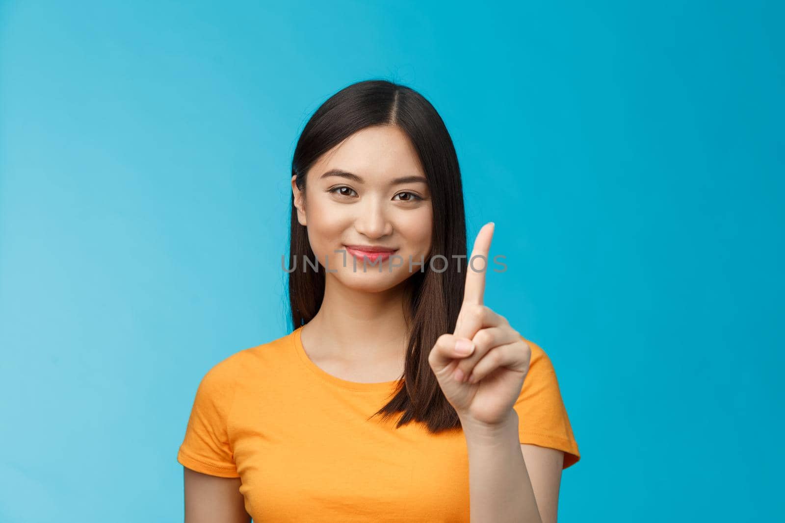 Close-up cheerful attractive tender asian woman with dark short haircut show number one, extend forefinger, smiling joyfully, friendly grin, stand blue background relaxed, casually make order.
