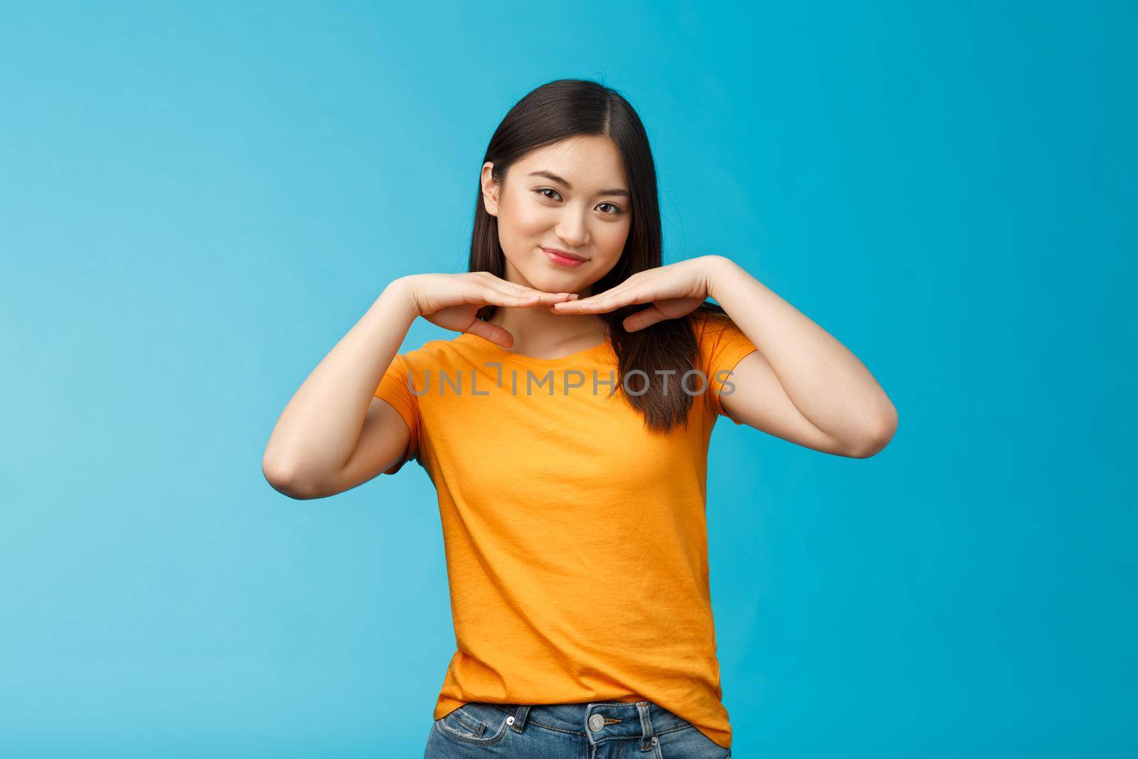Sweet lovely tender asian girl brunette, tilt head smiling show clean pure face hold palms under jawline satisfied after applying skincare product stand blue background wear yellow t-shirt.