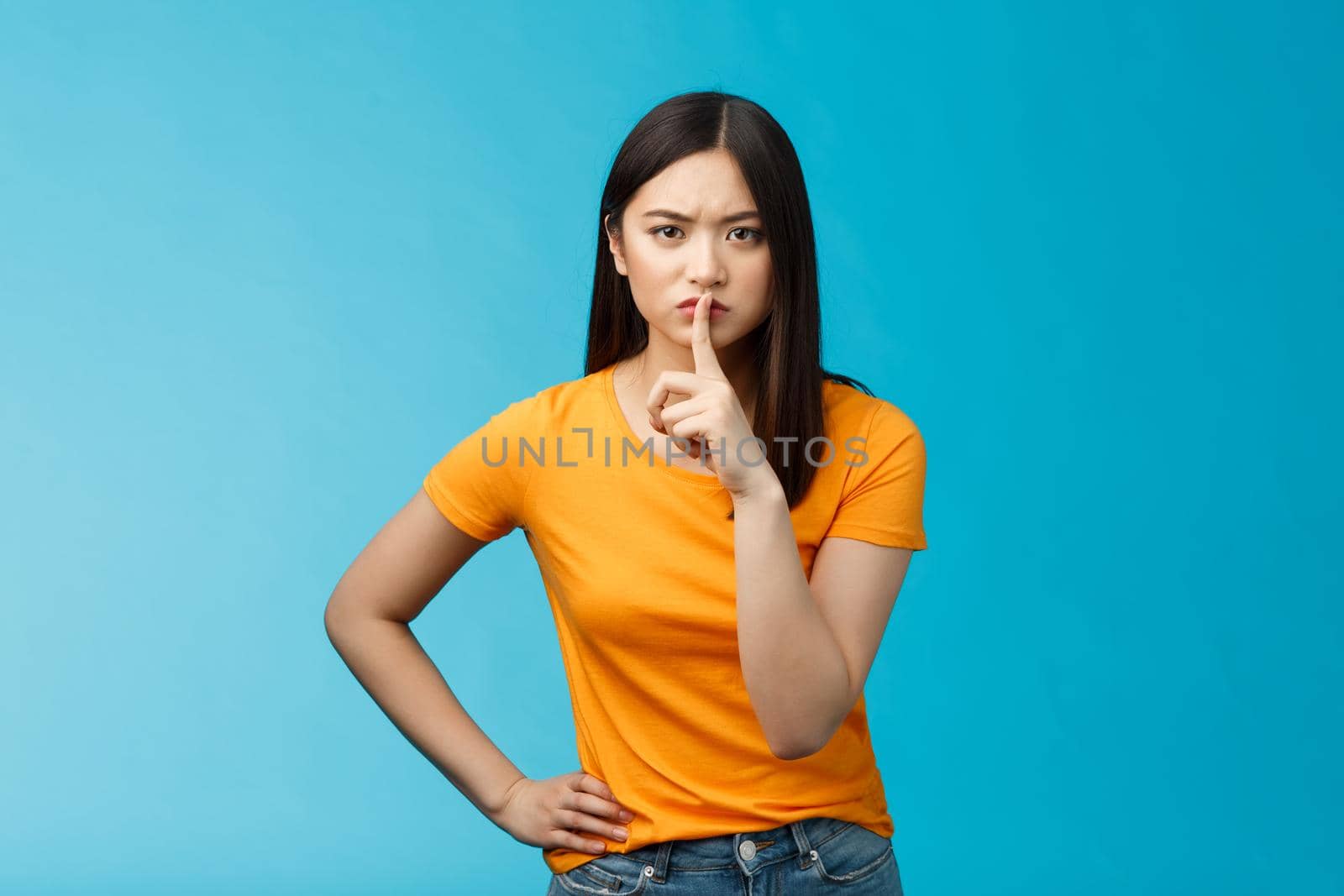 Hush keep quiet, behave well. Serious-looking strict asian elder sister shushing bending towards camera frowning, hold hand waist encouraged, asking not share secret, stand blue background.