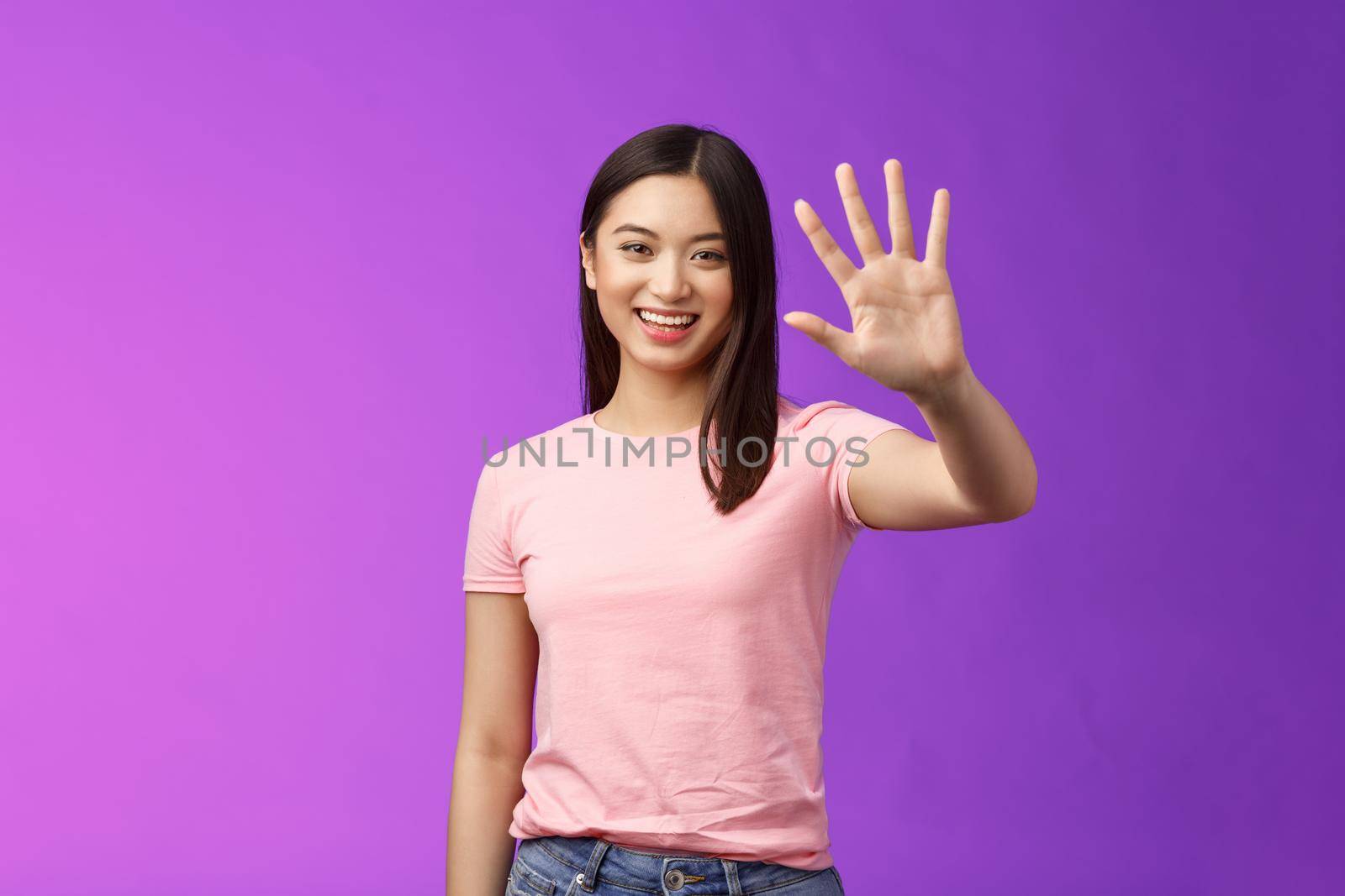 Friendly cute asian tender girl raise palm waving goodbye, smiling joyfully, farewell, gladly welcome friends inviting come in, saying hi hello, greeting, stand purple background. Copy space