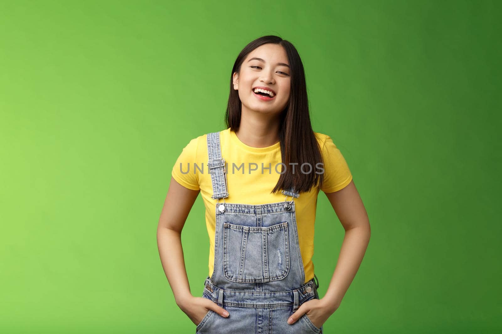Carefree enthsuastic outgoing attractive teenage asian girl having fun laughing friends, hold hands overalls pockets smiling toothy, take party easygoing conversation, stay positive green background by Benzoix