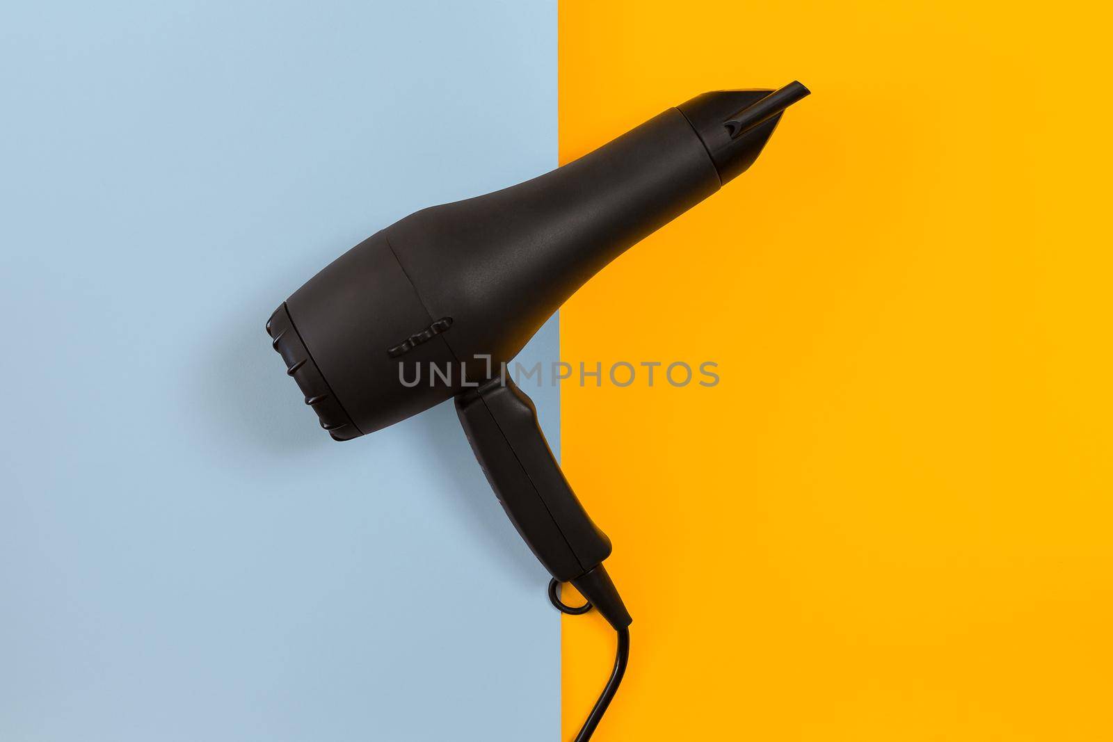 Black hair dryer on blue and yellow paper background. Top view. Copy space. Flat lay. Still life. Mockup