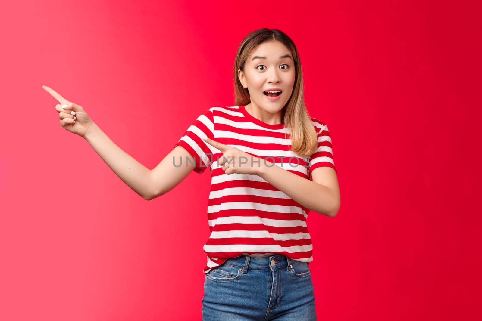 Hey hurry up look. Enthusiastic excited cute asian blond urban girl pointing left showing incredible offers, raise eyebrows thrilled talk camera smiling delighted stand red background impressed.