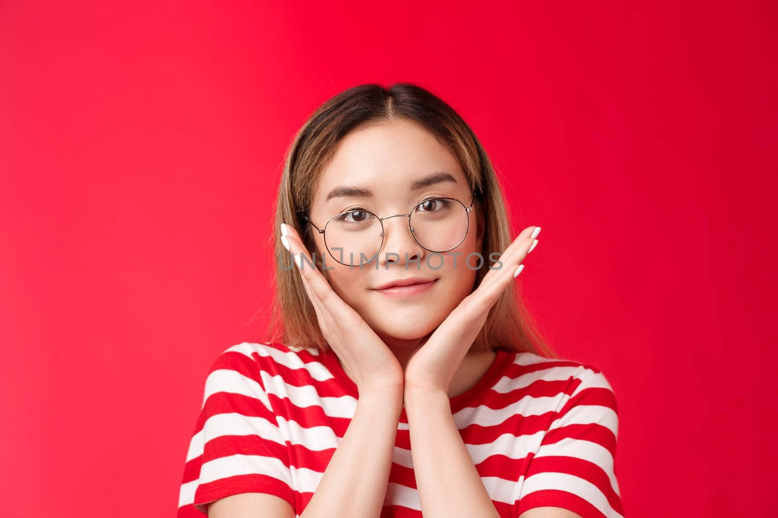Close-up tender cute asian teenage girl finally got rid pimples skin flaws, touch pure clean skin apply daily skincare products satisfied result, stand pleased red background smiling wear glasses.