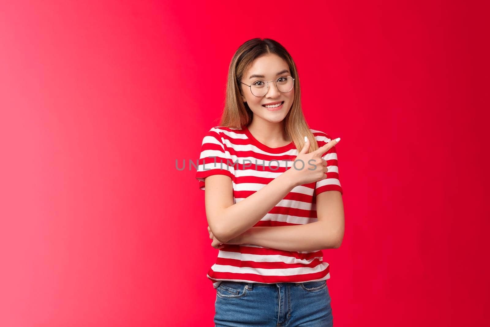 Cheerful friendly asian female blond hairstyle wear striped summer t-shirt glasses, pointing right showing you cool place smiling broadly, stand red background relaxed, introduce promo offer by Benzoix