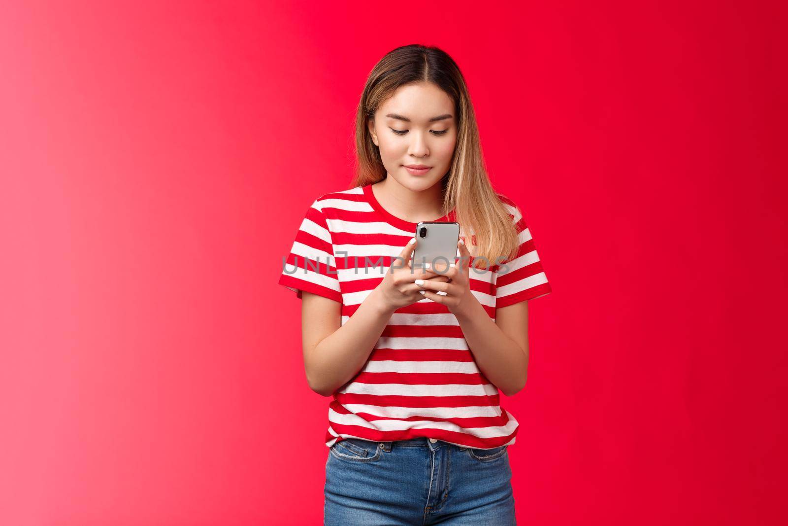 Popular asian female lifestyle blogger posting photo online hold smartphone look phone display scroll feed, woman smiling delighted relaxed using gadget stand red background by Benzoix
