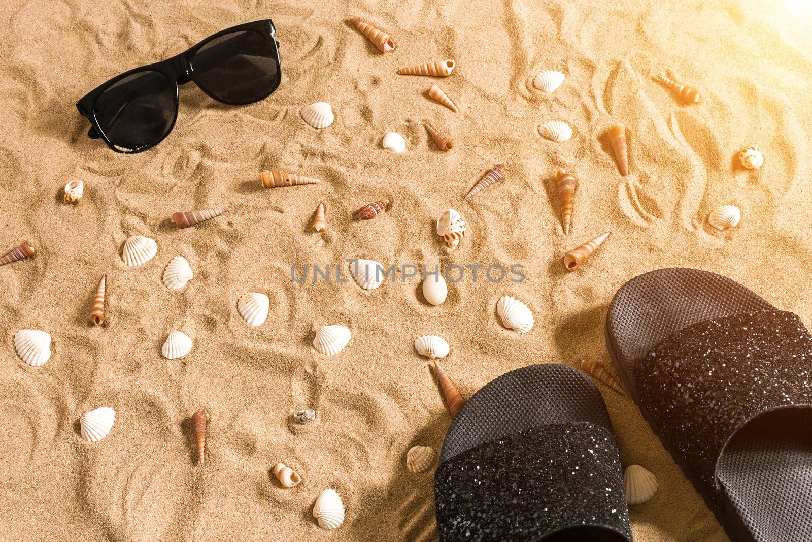 Black flip-flops and sunglass, seashell on sand. With place for your text. Top view. Copy space. Sun flare