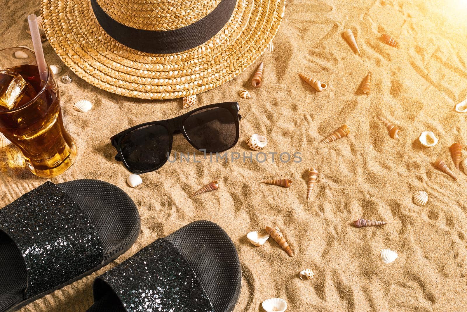 Summer beachwear, flip flops, hat, cold drink in a glass and seashells on sand beach. Top view. Copy space. Sun flare