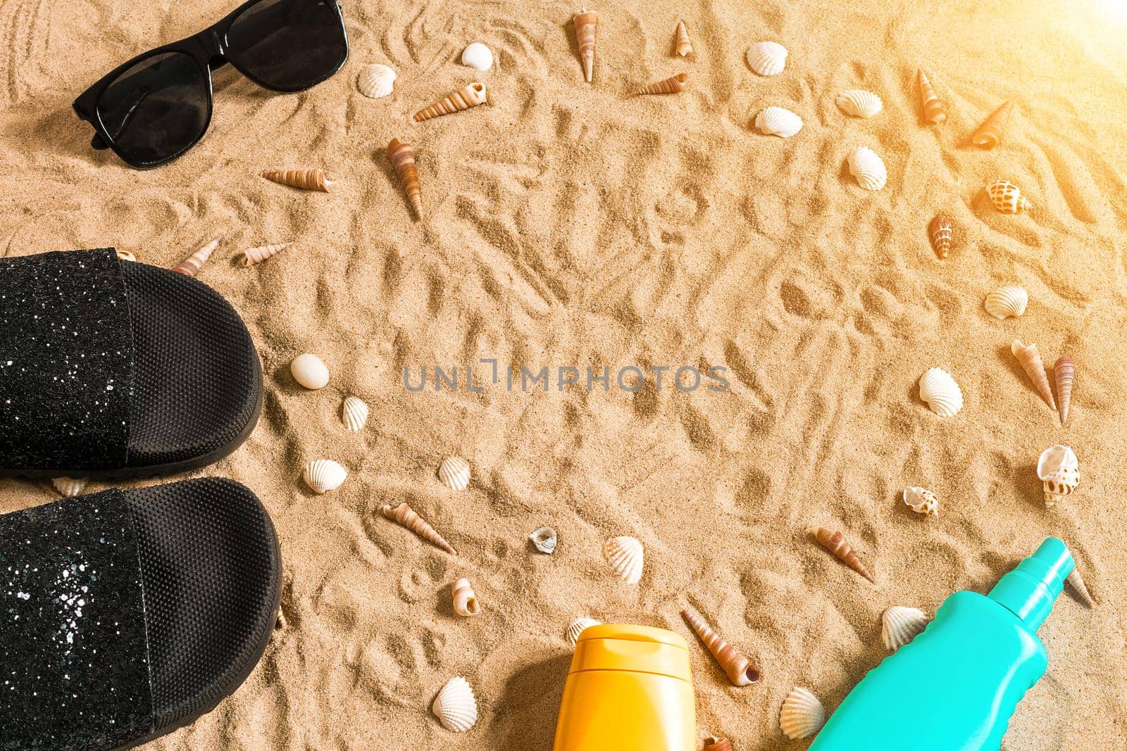 Black flip-flops and sunglass, seashell on sand. With place for your text. Top view. Copy space. Sun flare