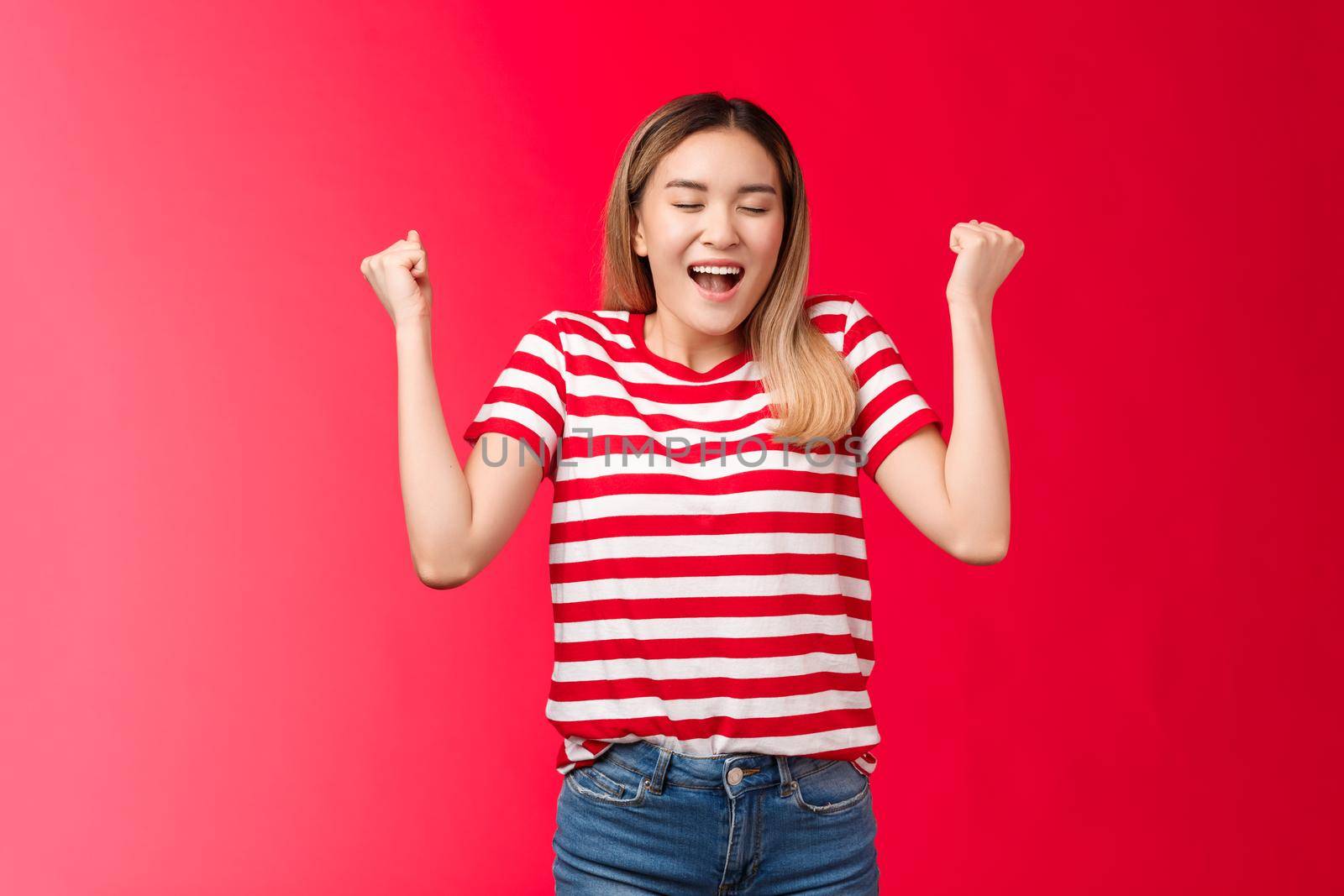 Hooray I did it. Triumphing cute cheerful asian blond girl make fist pumps closed eyes happy broad smile, celebrating accomplishment feel satisfaction finally winning achieve success red background.