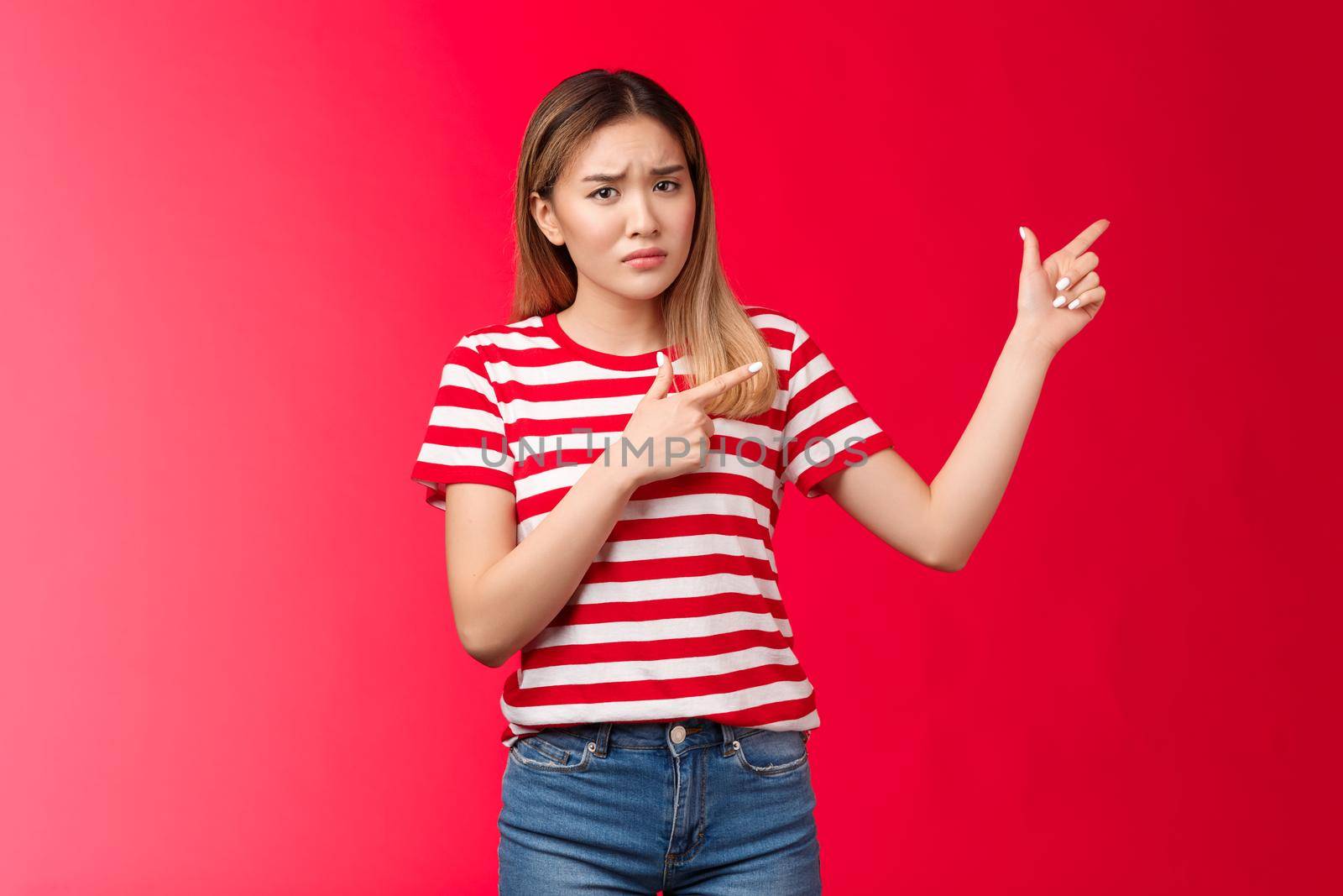 Displeased moody asian girlfriend acting childish immature pointing upper right corner upset, frowning doubtful unsatisfied, look camera hesitant and reluctant, stand unimpressed red background.