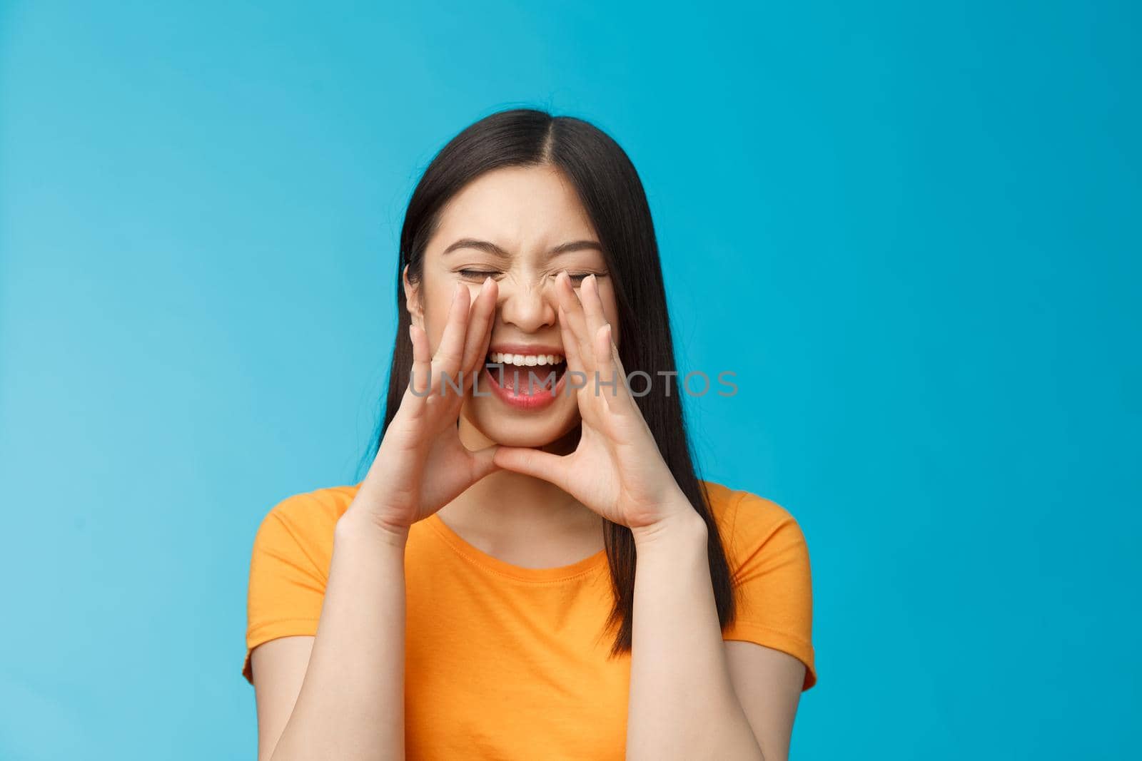 Carefree asian enthusiastic girl shouting long distance, calling friend, breaking free emotions, yelling out loud, hold hands near opene mouth rupor, searching someone in crowd, blue background.