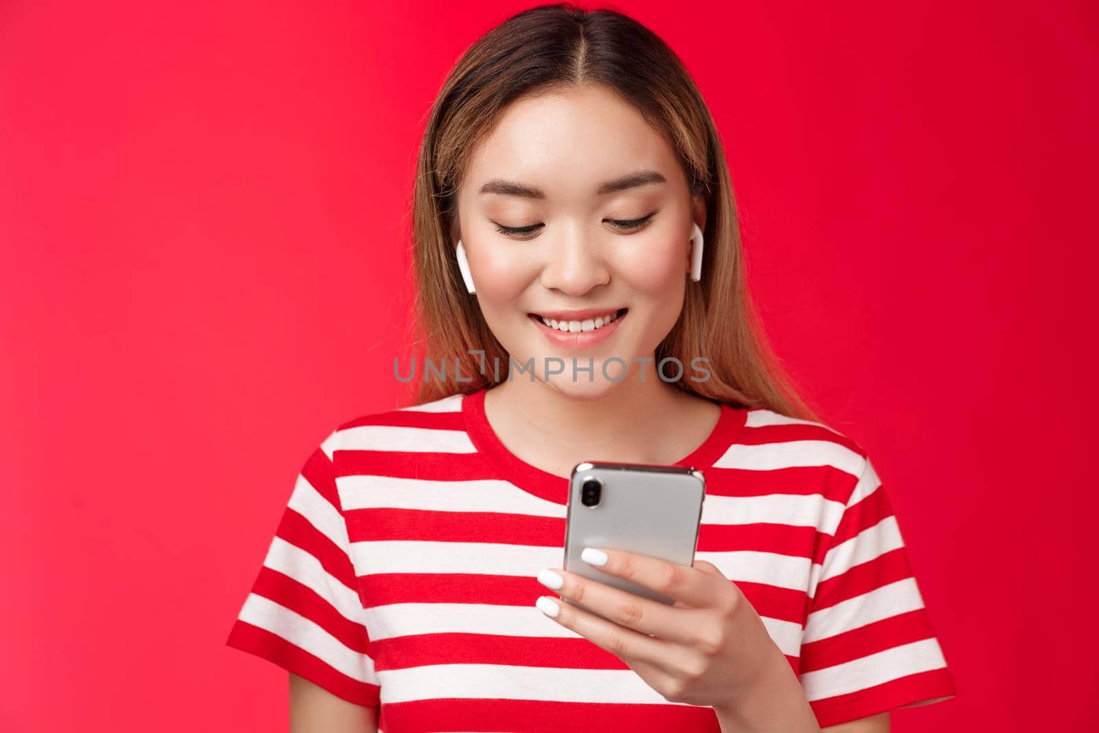Carefree modern asian blond girl picking song wear wireless earphones, listen music using online song platform app, hold smartphone look pleased smiling phone screen, stand red background.