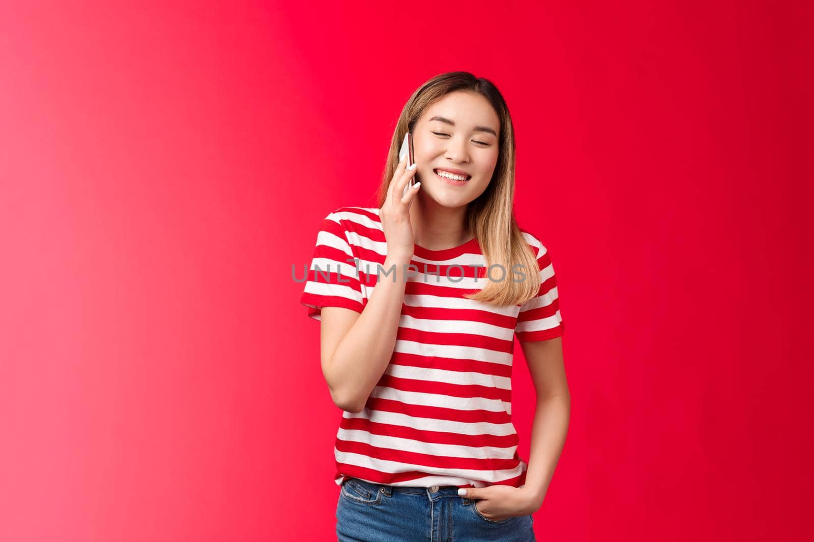 Carefree friendly good-looking blond asian girl having funny conversation, talking phone, close eyes joyfully smiling calling friend hold smartphone near ear, laughing over hilarious voice message.