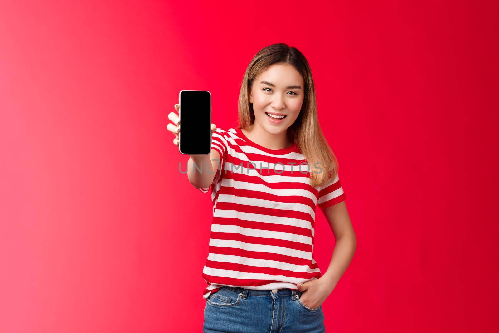 Check out my game score. Carefree good-looking blond asian woman extend arm showing smartphone screen smiling broadly stand relaxed introduce cool app, bragging bank account red background.