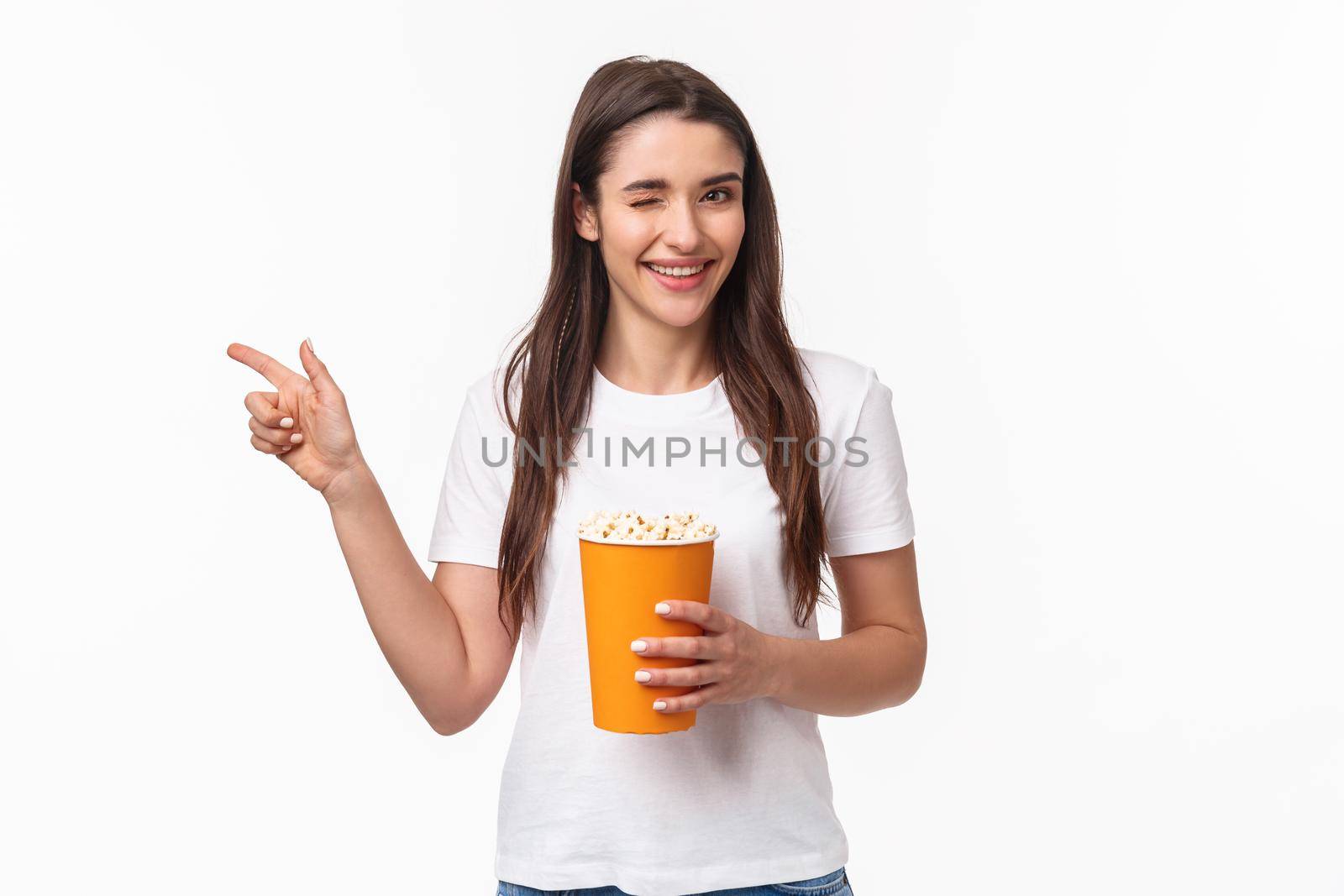 Entertainment, fun and holidays concept. Portrait of cheeky cute young girl in t-shirt, wink and smiling, pointing finger left inviting come and watch movie together in cinema, hold popcorn.