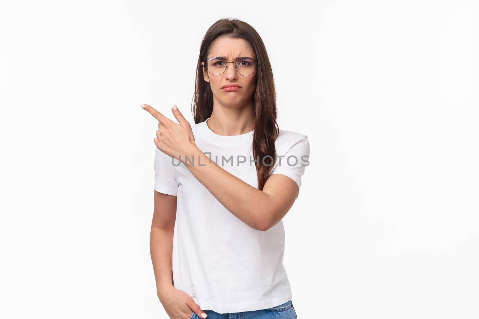 Waist-up portrait of moody, disappointed and complaining cute brunette woman in glasses, frowning and grimacing, sulking defensive, pointing finger upper left corner at something offensive.