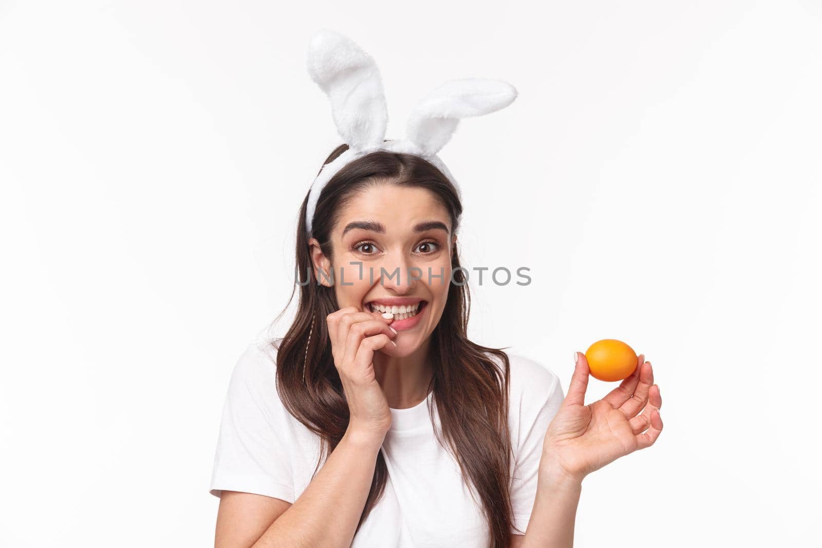 Easter, holidays and spring concept. Close-up portrait of silly and cute, happy giggling girl in rabbit ears, made her first colored egg on Easter day, biting finger, looking camera joyful.