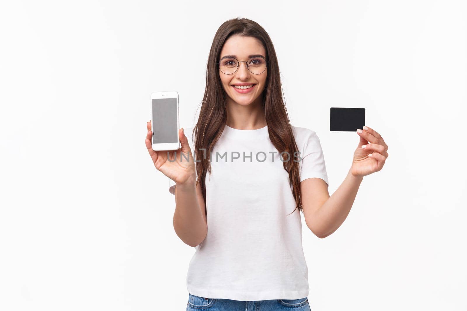Portrait of happy friendly-looking young woman in t-shirt and glasses, showing credit card and mobile phone screen as advertise online application, internet purchase, non-cash payment.