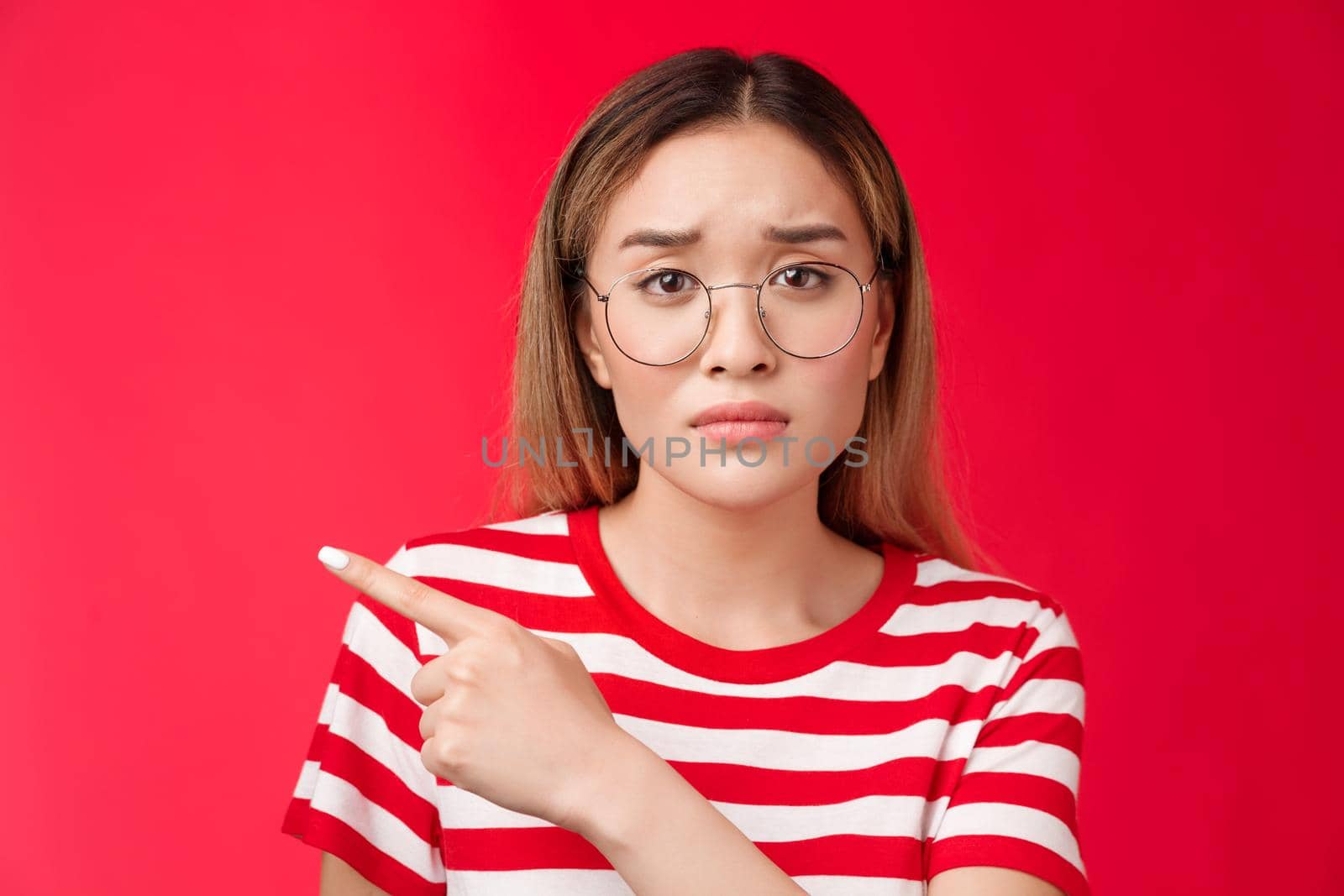 Sad cute asian female coworker tired working wear glasses frowning, pointing left look camera upset drained feel pressured distressed, stand red background unhappy wear striped t-shirt.