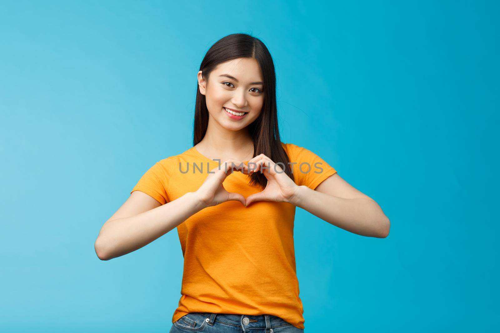 Lovely caring attractive asian girlfriend show heart sign cherish and value relationship, smiling broadly supportive, stand blue background wear yellow t-shirt, promote peace and happiness by Benzoix