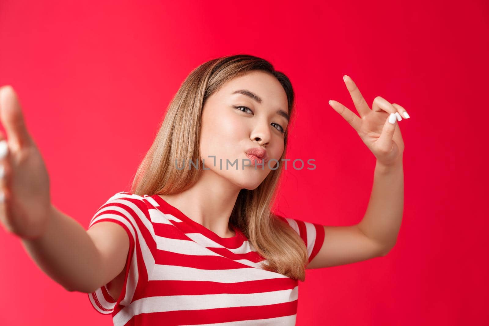 Close-up friendly outgoing cute asian girl blond hairstyle taking photograph, hold smartphone fold lips silly kiss expression, show peace victory sign, make selfie smartphone red background.