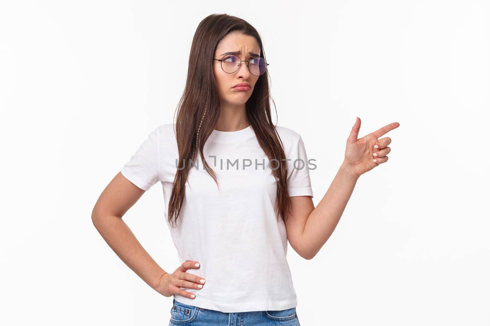 Waist-up portrait of uneasy, sad and gloomy young brunette woman in glasses, looking and pointing finger right with sulking unhappy face, express jealousy or regret, grieving white background.
