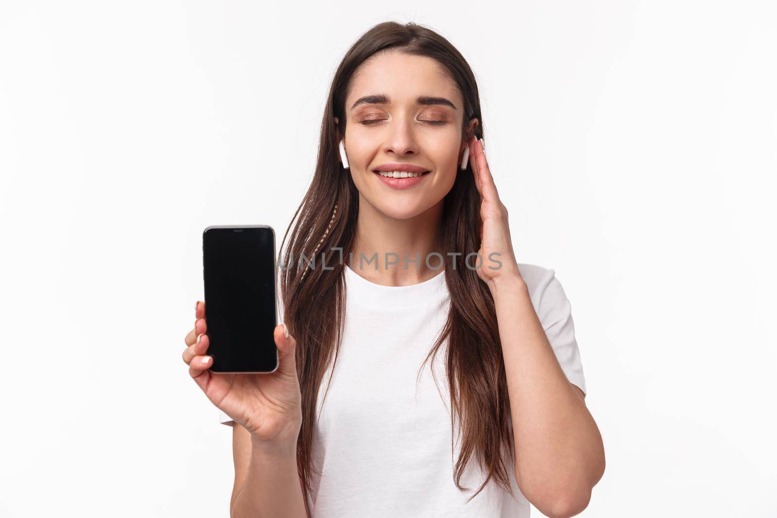 Close-up portrait of delighted, romantic young woman feeling satisfaction of listening favorite playlist, showing mobile phone screen, close eyes smiling and wearing wireless headphones.