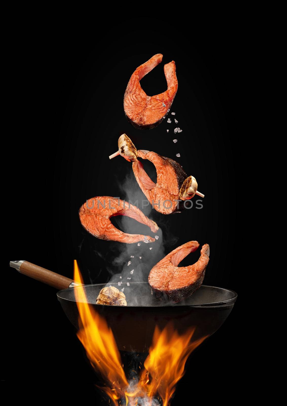 Wok pan with smoke above fire is frying salmon steaks with garlic against black studio background. Cooking concept. Close up, copy space