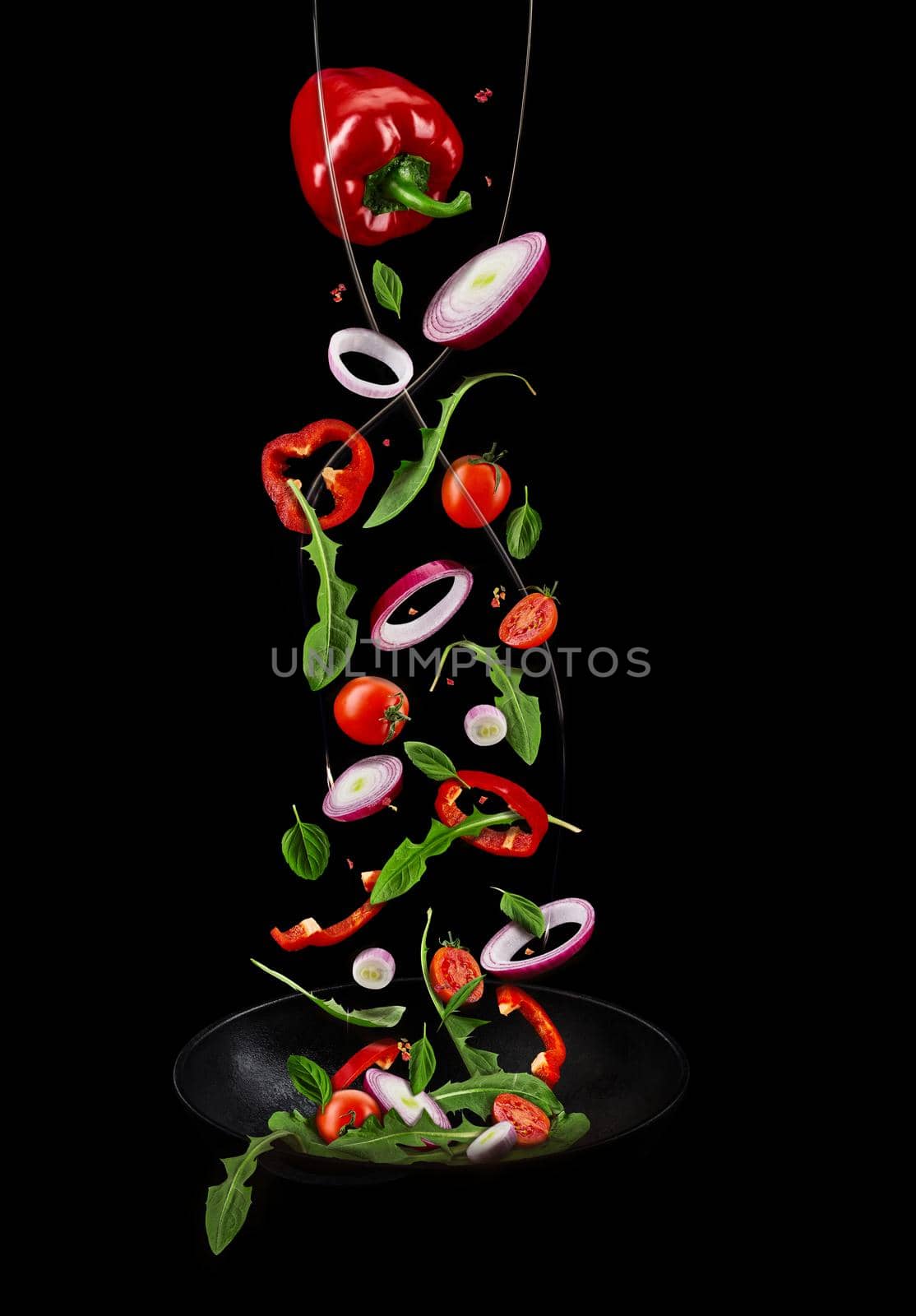 Wok pan, falling ripe cherry tomatoes, red bell pepper, onion, green basil and arugula against black studio background. Poured by a water stream. Cooking concept. Close up, copy space