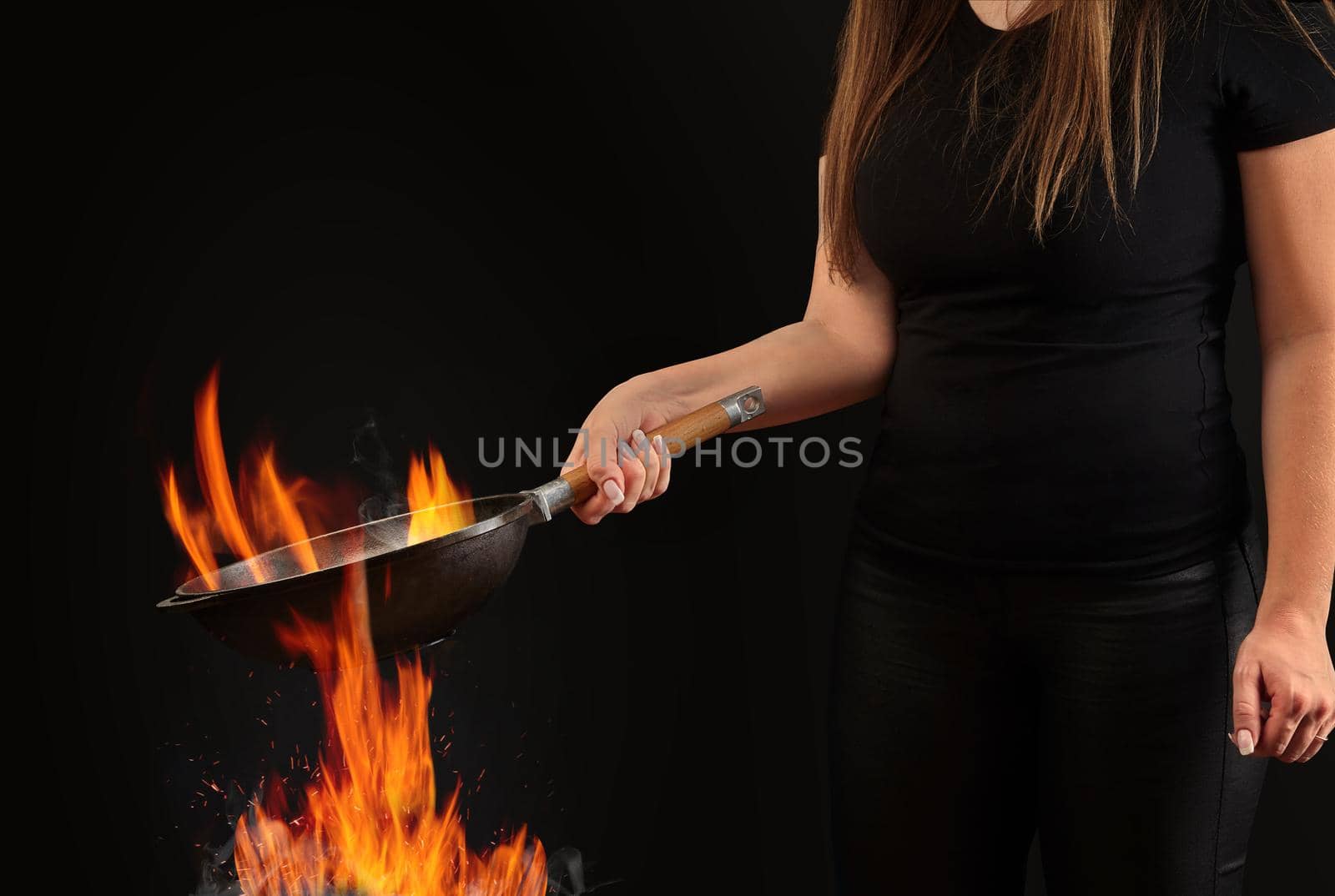 Brunette lady with tattooed hands and long hair, dressed in leggings and t-shirt. Holding a wok pan above fire against black studio background. Cooking concept. Close up, copy space, side view