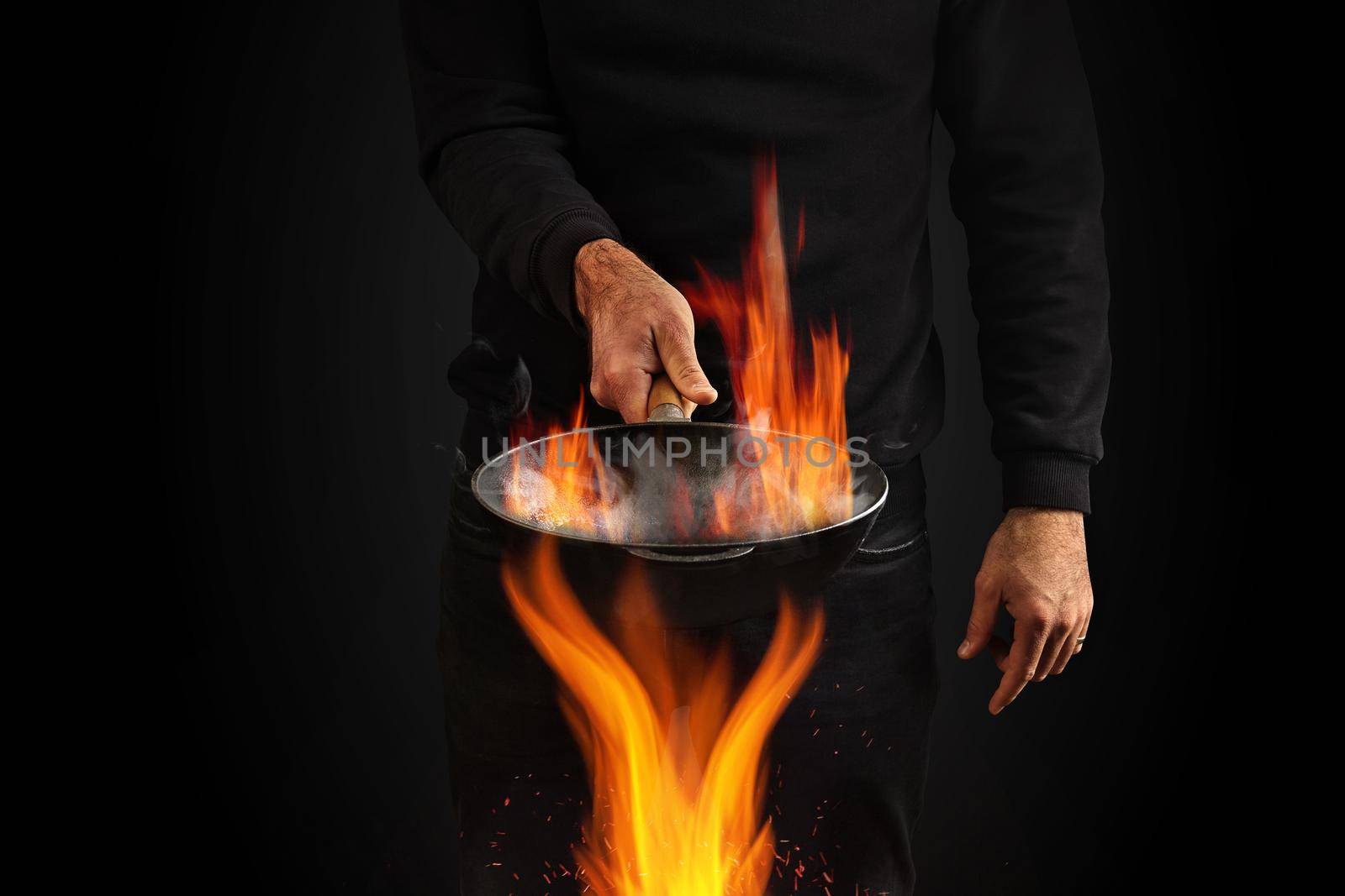 Young man dressed in jumper and jeans. He is holding burning wok pan with smoke above fire, against black studio background. Cooking concept. Close up, copy space