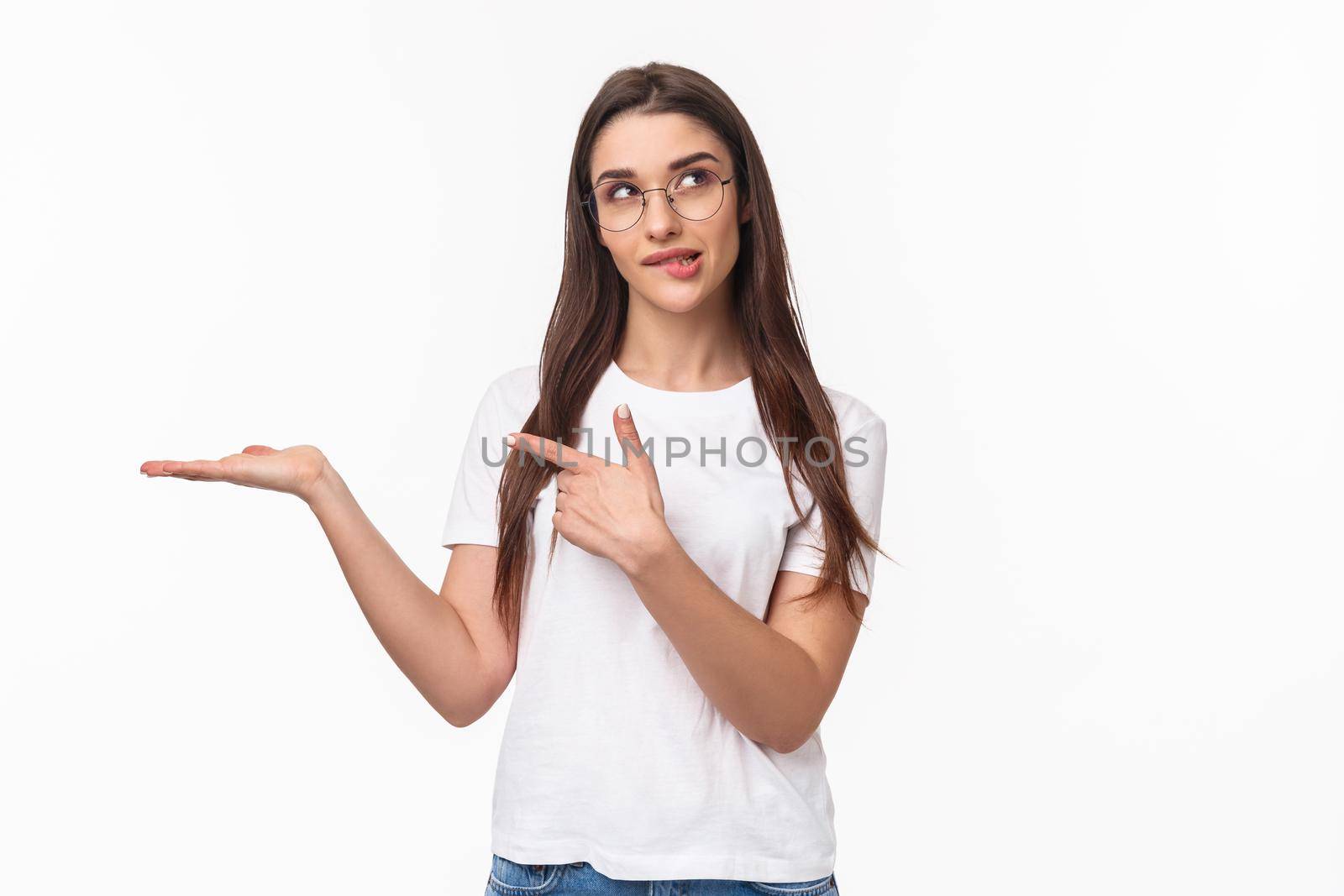 Waist-up portrait of thoughtful, pretty young woman holding arm as if have something lying in it, pointing at product with pondering face, bite lip look up thinking, white background.