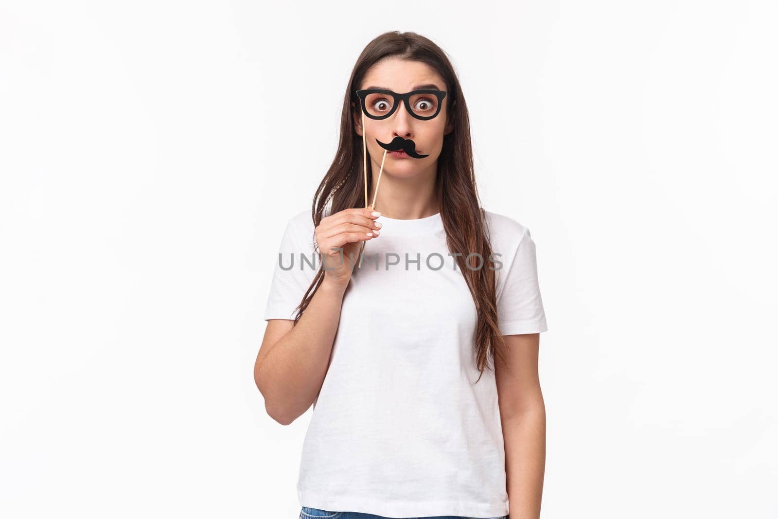 Entertainment, fun and holidays concept. Portrait of funny, playful and carefree girl holding carnaval mask sticks, fancy moustache and glasses, staring camera with popped eyes, white background.