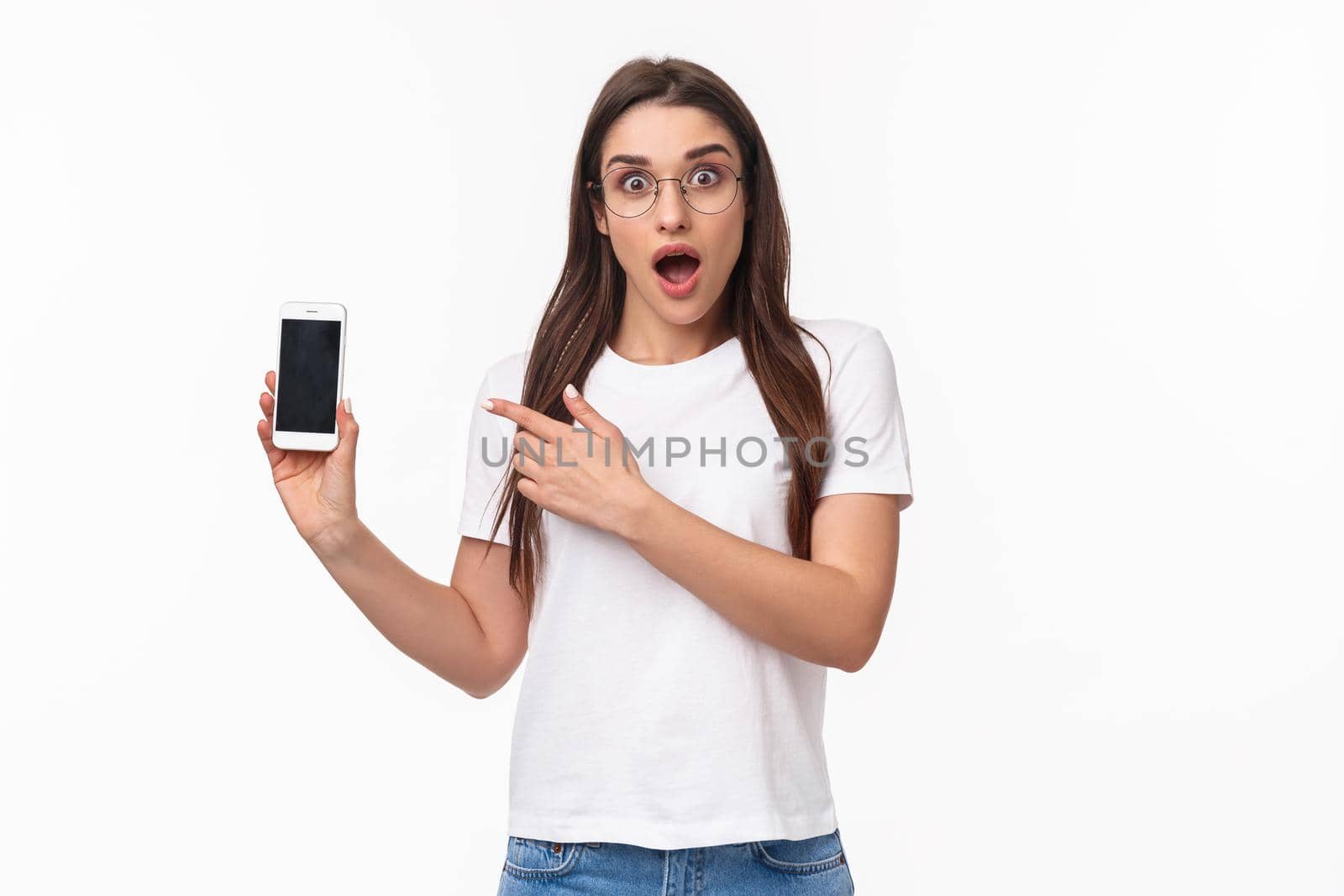 Communication, technology and lifestyle concept. Portrait of impressed and excited young brunette girl in glasses, pointing at mobile phone display with gasp and amazed face, white background by Benzoix