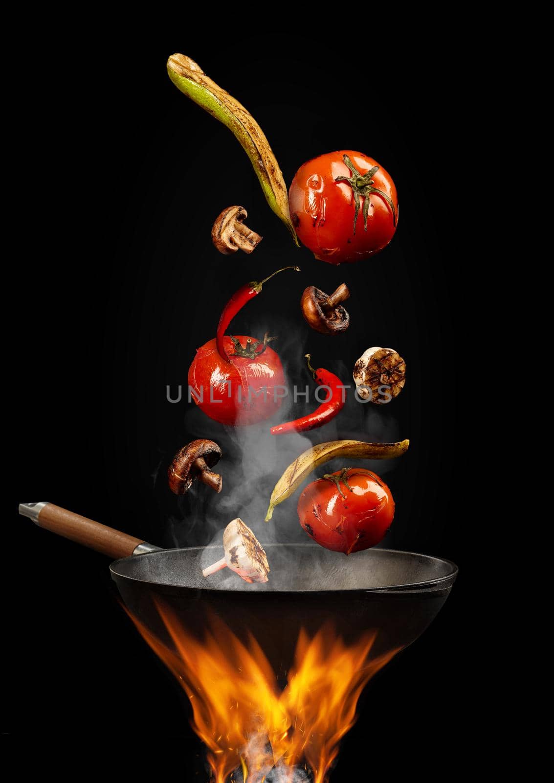 Wok pan with smoke above fire is frying red tomatoes, hot pepper, mushrooms, zucchini, garlic against black studio background. Close up by nazarovsergey