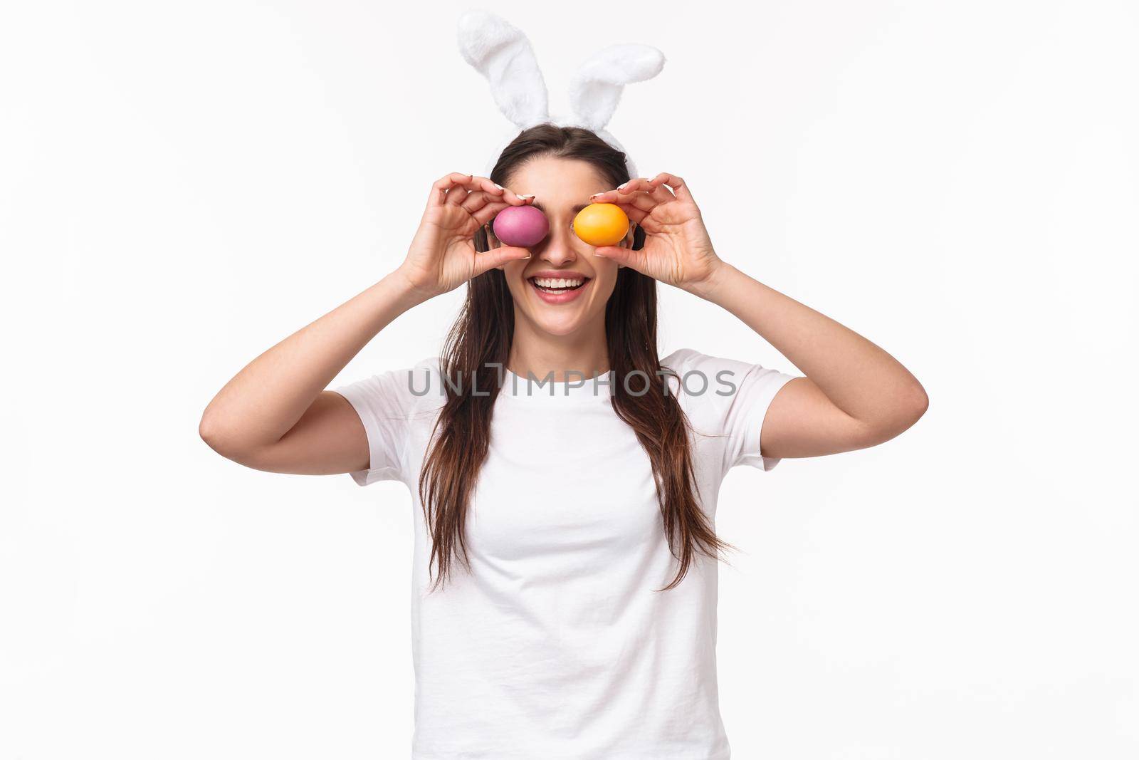 Portrait of funny and carefree cute young girl in rabbit ears, making eyes from colored eggs, celebrating Easter day, having fun, being in playful mood entertain kids on holiday, white background.