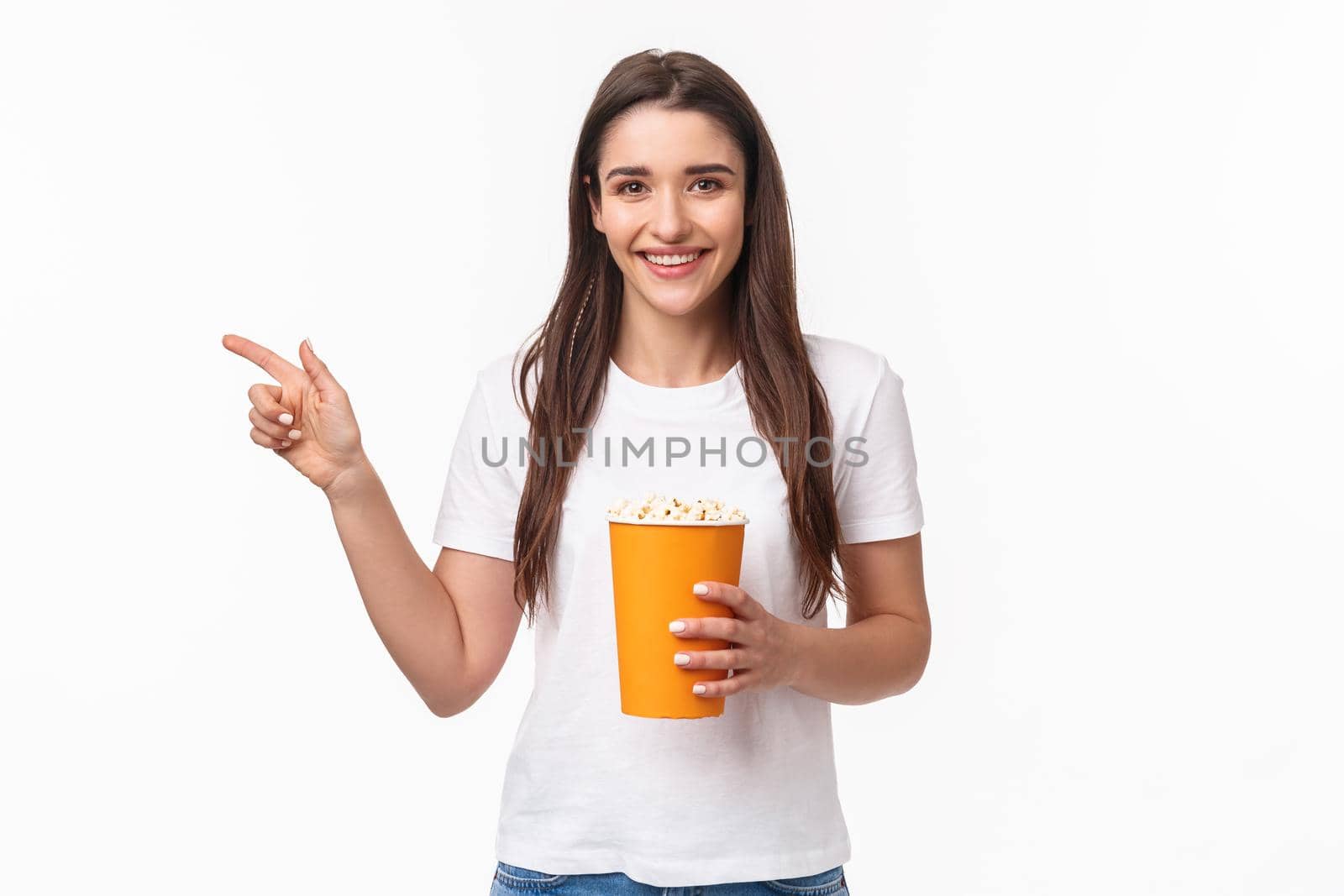 Entertainment, fun and holidays concept. Portrait of cheerful cute young girl waiting for friend in cinema, movie starting soon, pointing finger left inviting come join her, smiling, hold popcorn by Benzoix