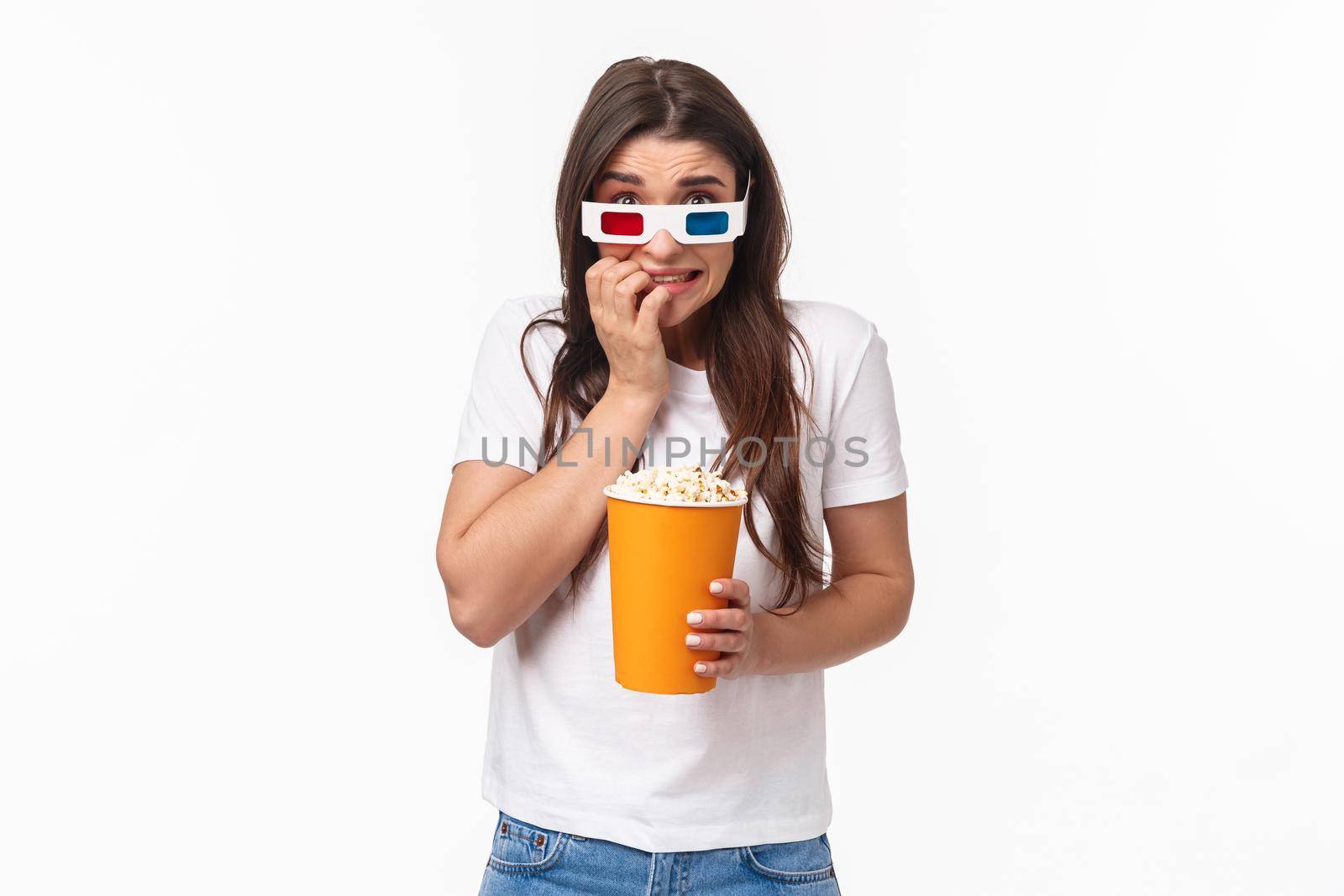 Entertainment, fun and holidays concept. Portrait of scared young cute girl in 3d glasses, eating popcorn, stooping and biting fingers as being scared of horror movie watching in cinema by Benzoix