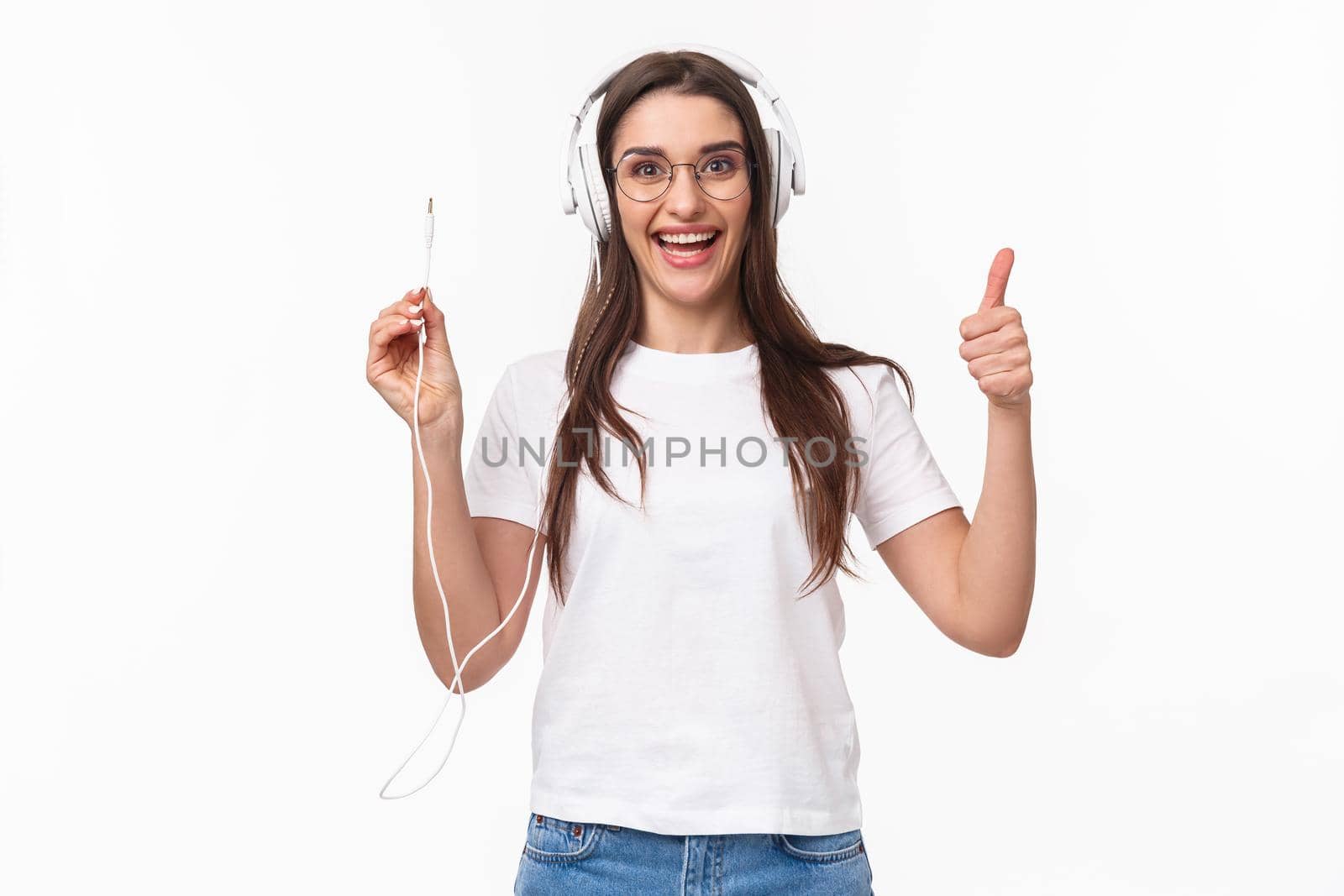 Waist-up portrait of excited, smiling happy and enthusiastic young girl in headphones, holding wire of earphones and show thumbs-up, ready to plug in to smartphone and listen some music.