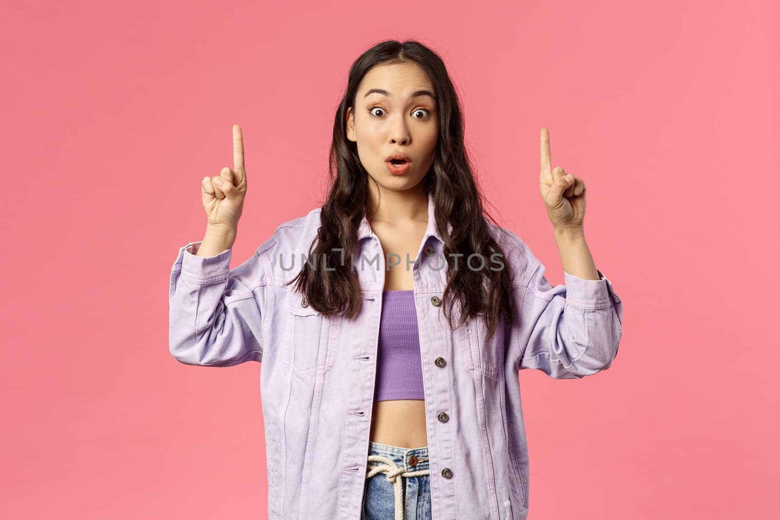 Portrait of impressed and surprised beautiful young girl in denim jacket, crop-top, pointing fingers up and staring at camera startled, gasping amazed, showing something cool, pink background.