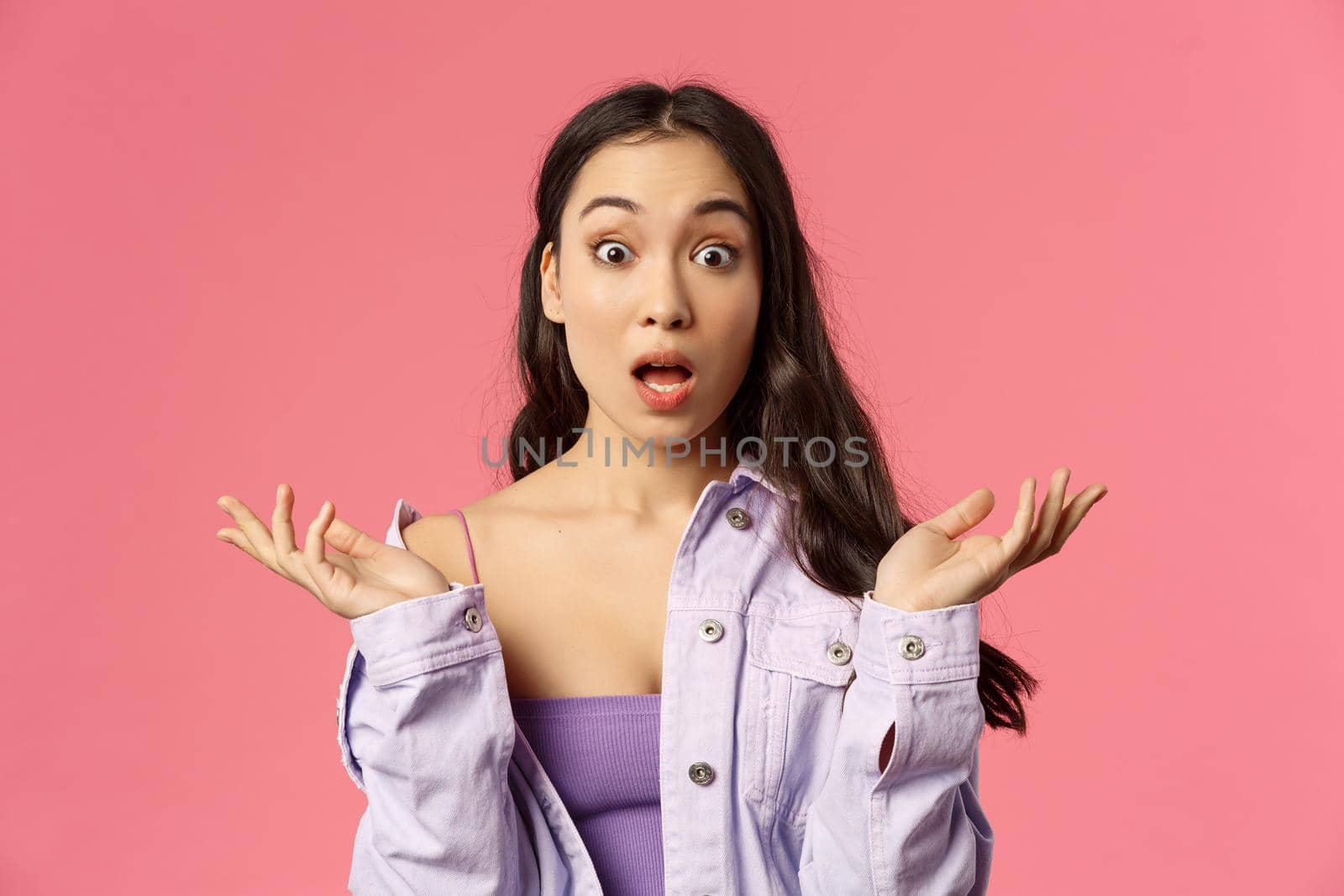 Close-up portrait of speechless, impressed young asian woman, spread hands sideways, gasping and open mouth wide from amazement, see something incredible, pink background.