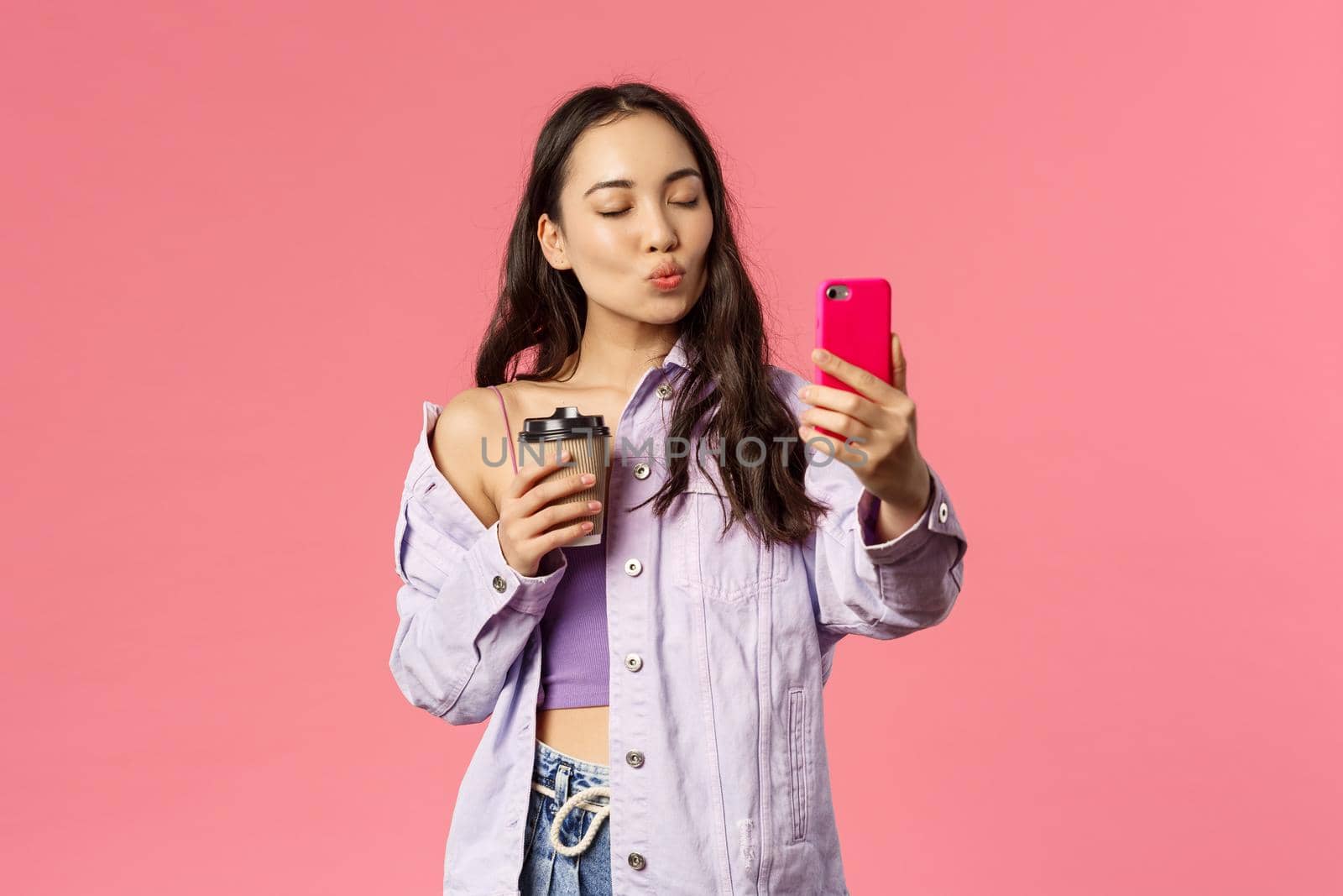 Portrait of stylish good-looking female blogger, lifestyle internet influencer taking selfie with take-away coffee from her favorite cafe, hold mobile phone, pouting for kiss and close eyes.